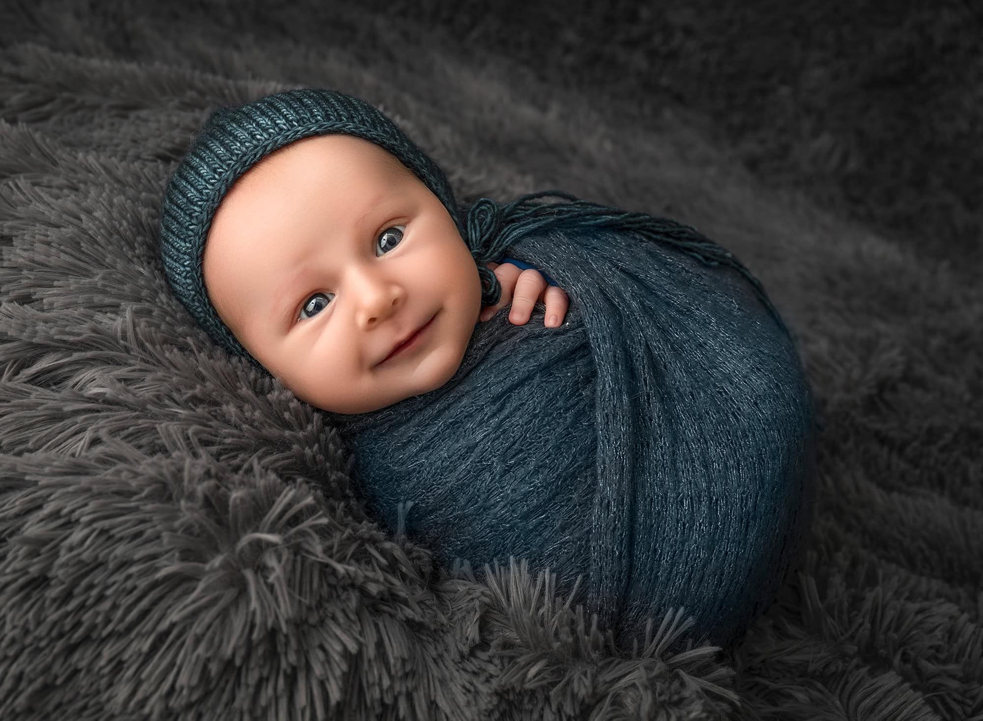 Levi in teal wrap and bonnet, smiling with bright eyes on a textured gray blanket.