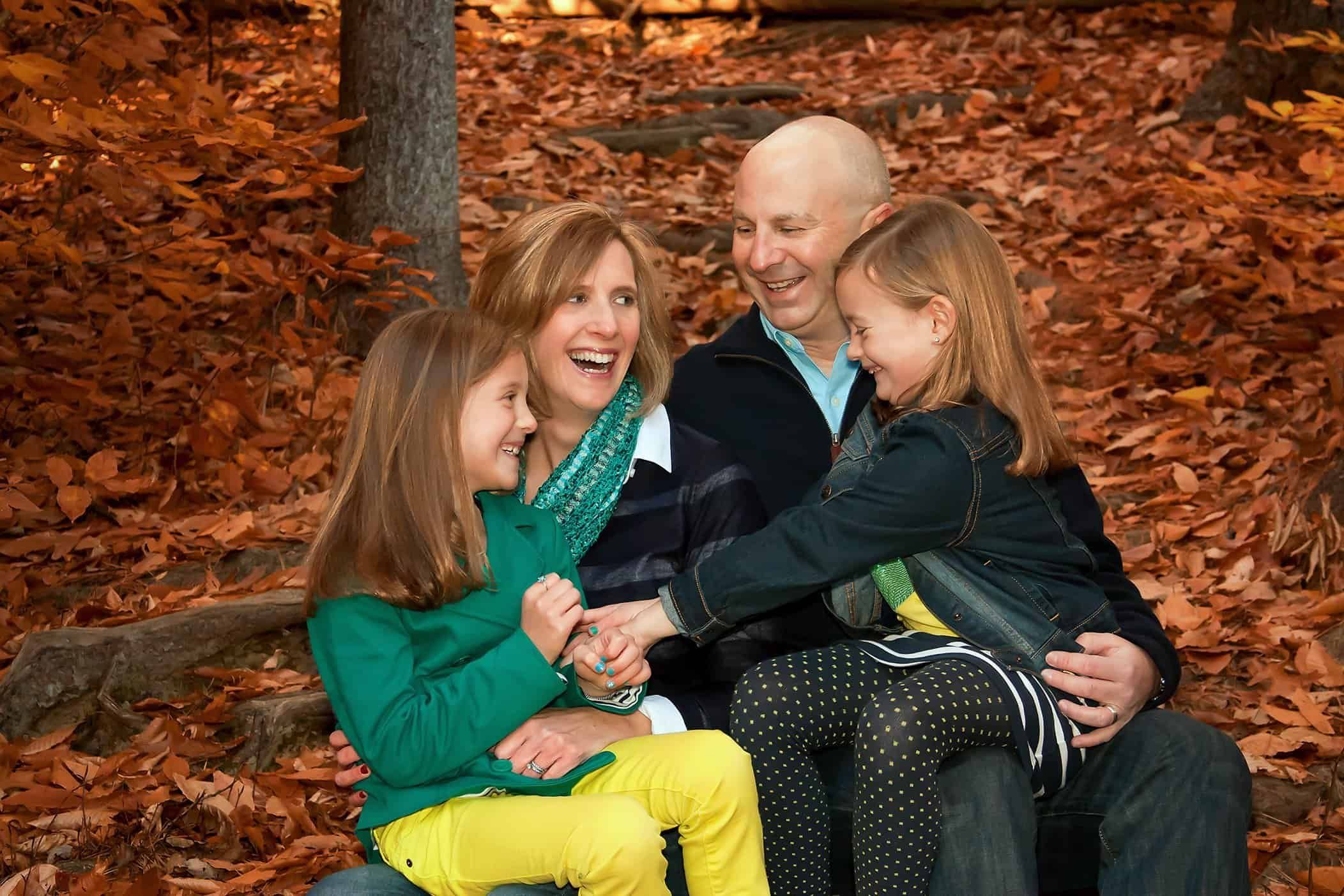 Mom, Dad and twin 7 year old girls sitting in orange fall leaves and laughing together