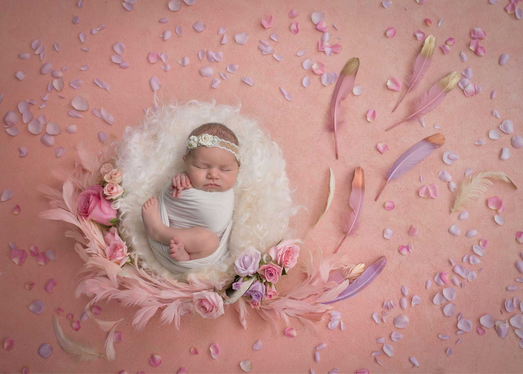 newborn baby girl sleeping in cream fluff on pink background with feathers and flowers