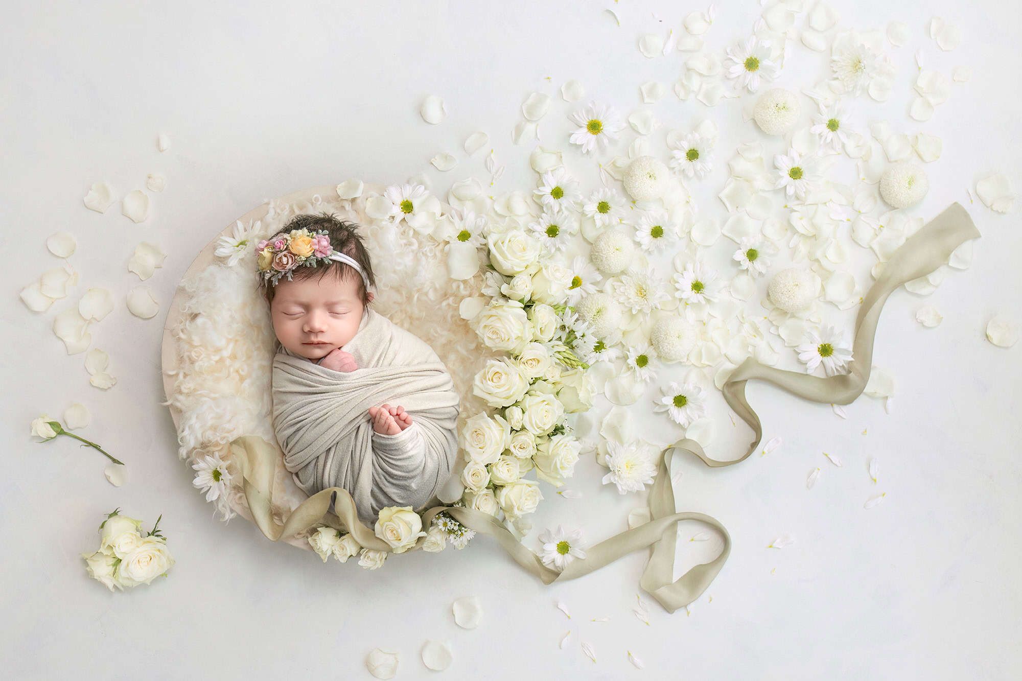 Swaddled newborn in cream wrap surrounded by white roses and ribbon.