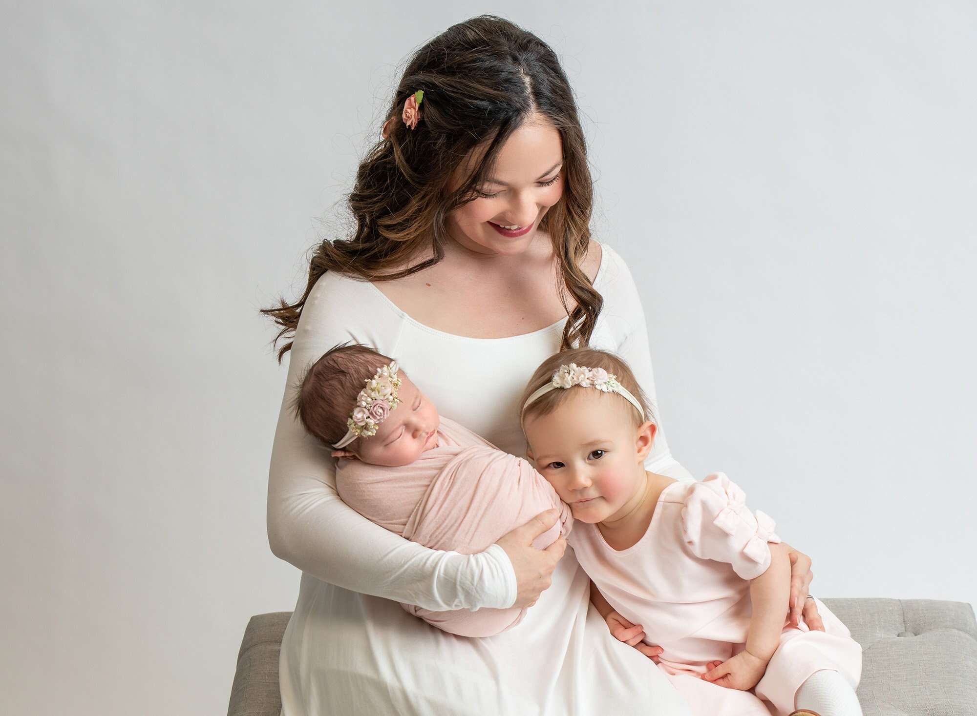 mom in white dress sitting on gray couch holding daughters dressed in pink