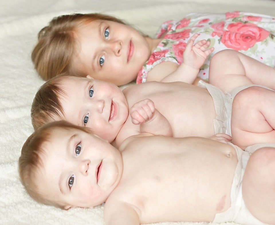 Fraternal twins and their big sister lying side by side with all of their eyes looking at you