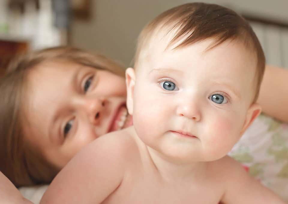Baby boy with blue eyes looking straight ahead with his big sister peeking out behind him