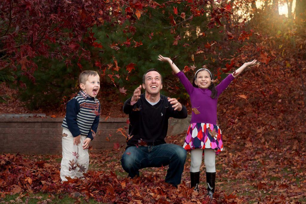 Daddy and two kids throwing orange fall leaves into the air with the setting sun in the background