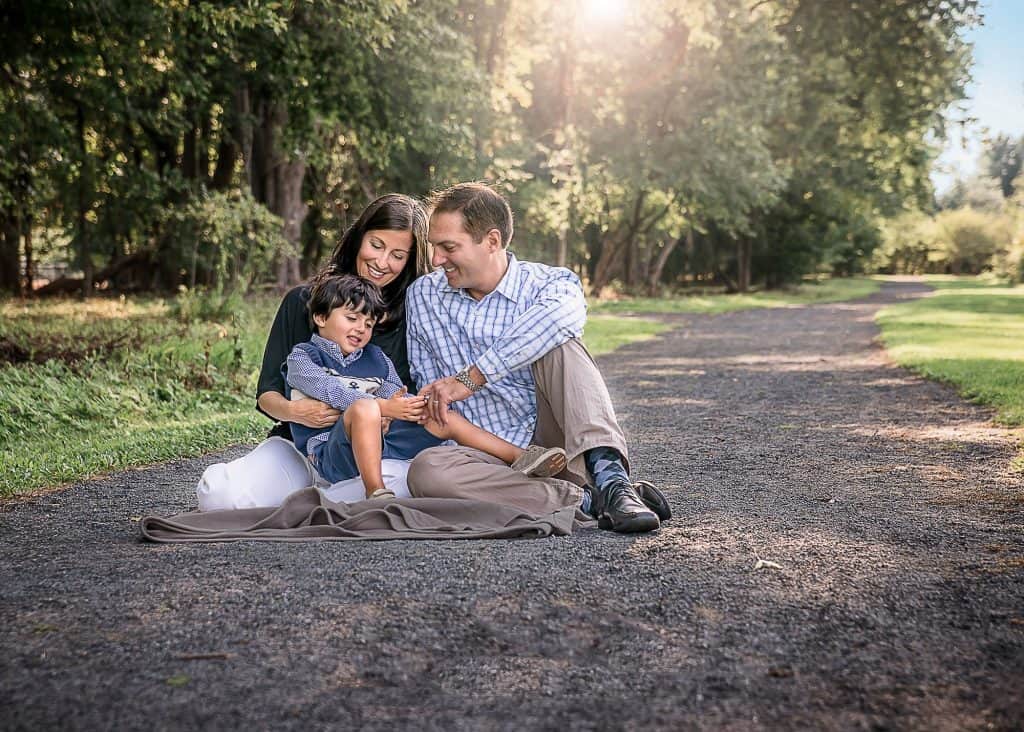 Family of 3 sitting on a blanket outside in summer with the sun setting behind them