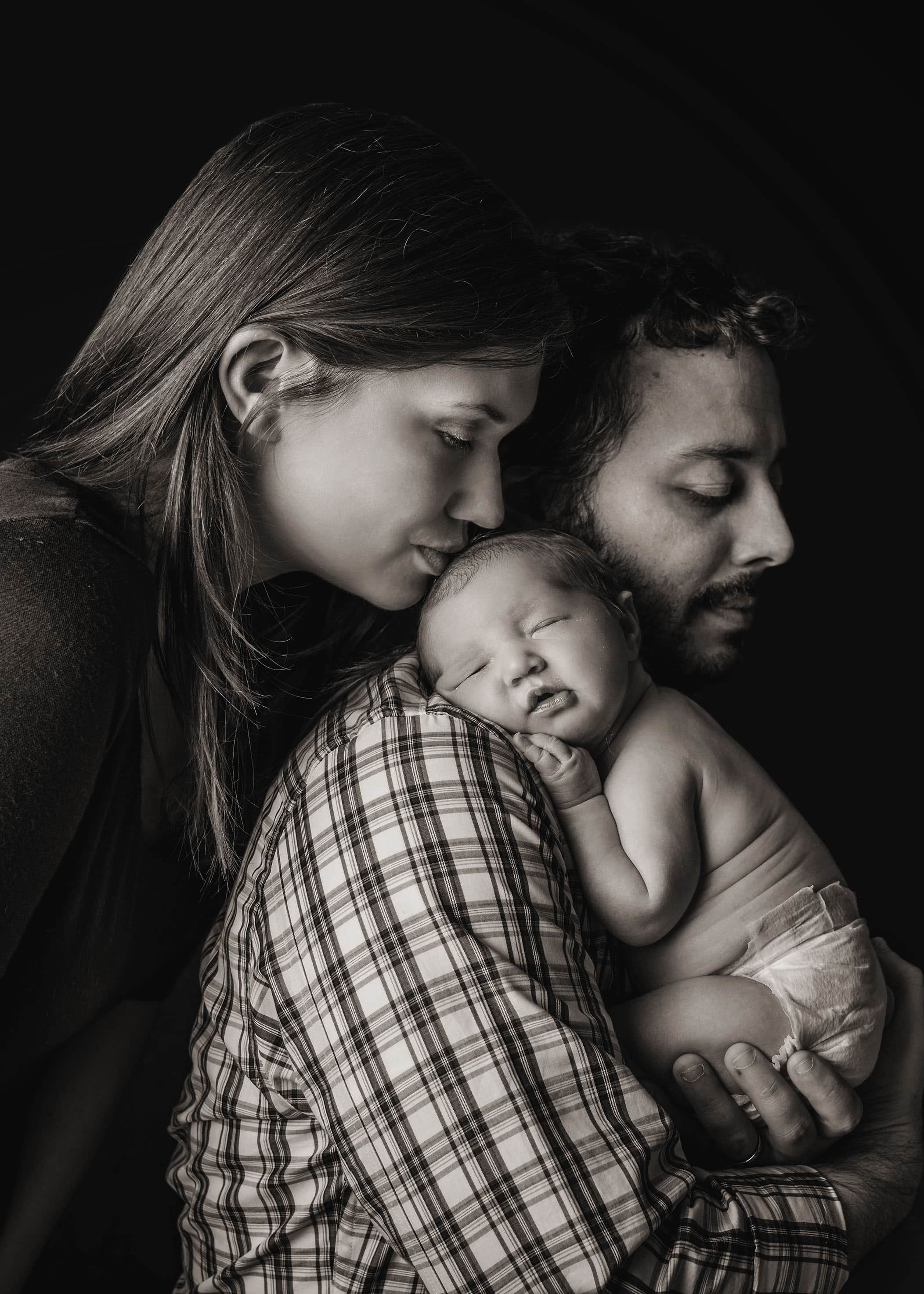 Dad holding newborn baby on his shoulder while mom kisses her head One Big Happy Photo