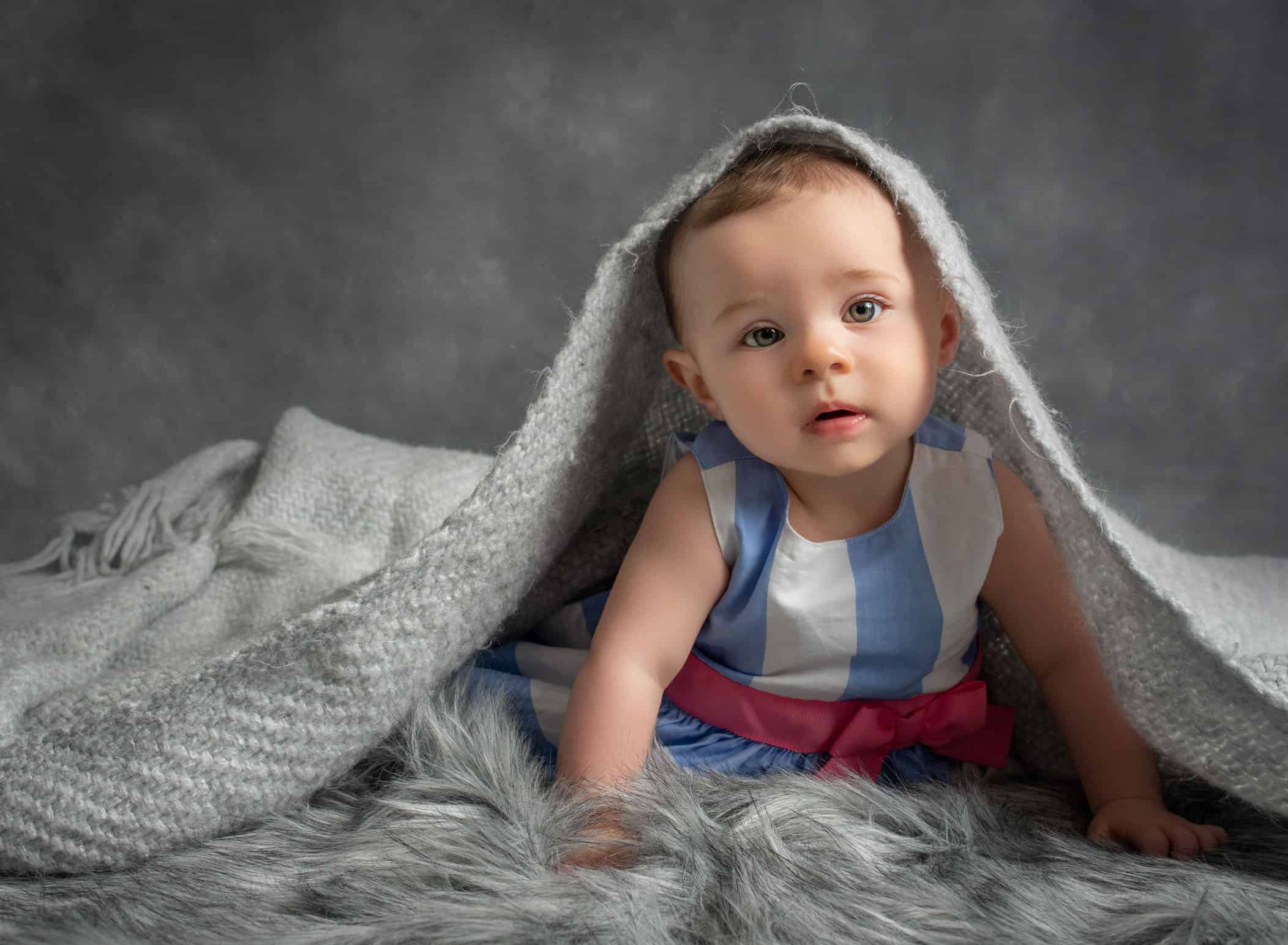 6 month old baby girl being serious under the edge of a grey blanket on grey fur rug