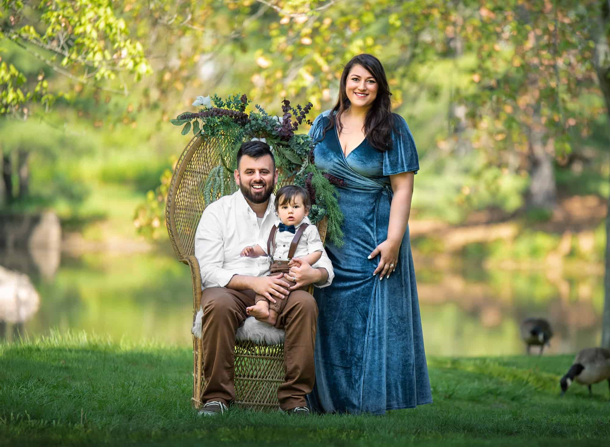 dad holding one year old boy sitting in his lap while sitting on a wicker chair in nature dressed up in greenery next to his wife