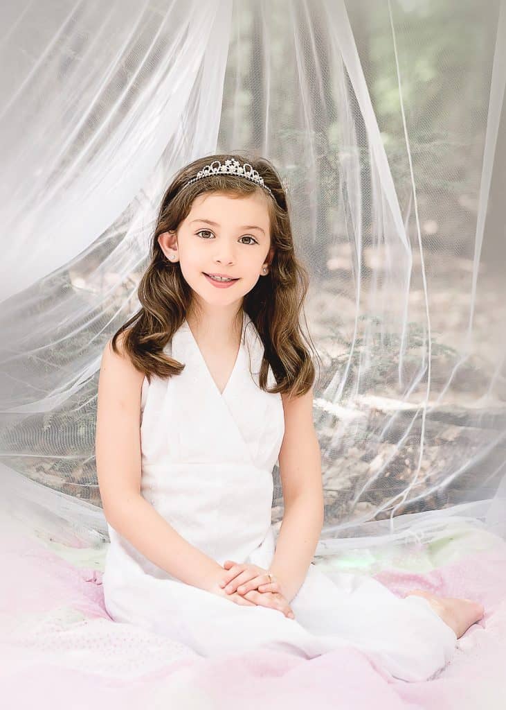 little brown haired girl sitting in a white mosquito net in the woods