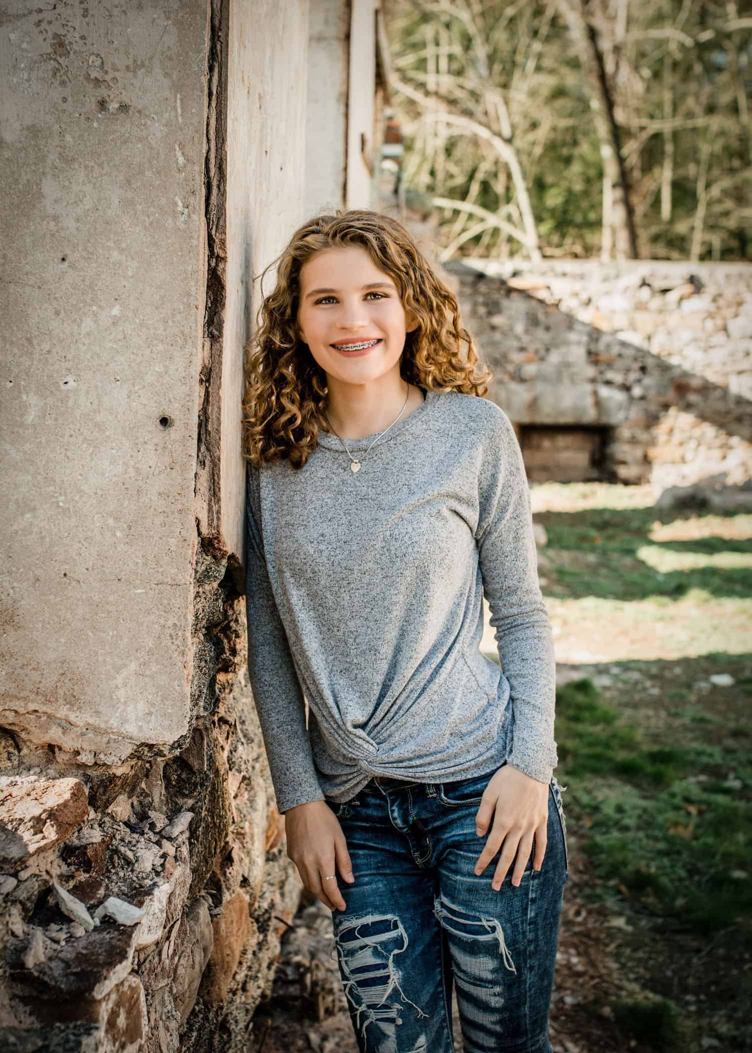 Teenage girl leans against wall of building ruins outside smiling