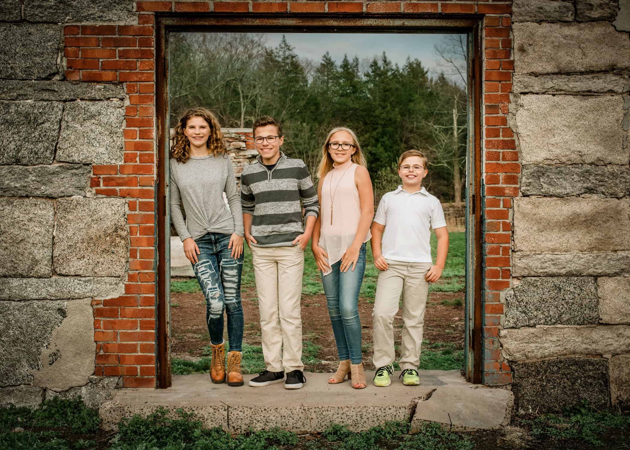 Four young siblings stand in old doorway of building ruins outside at park