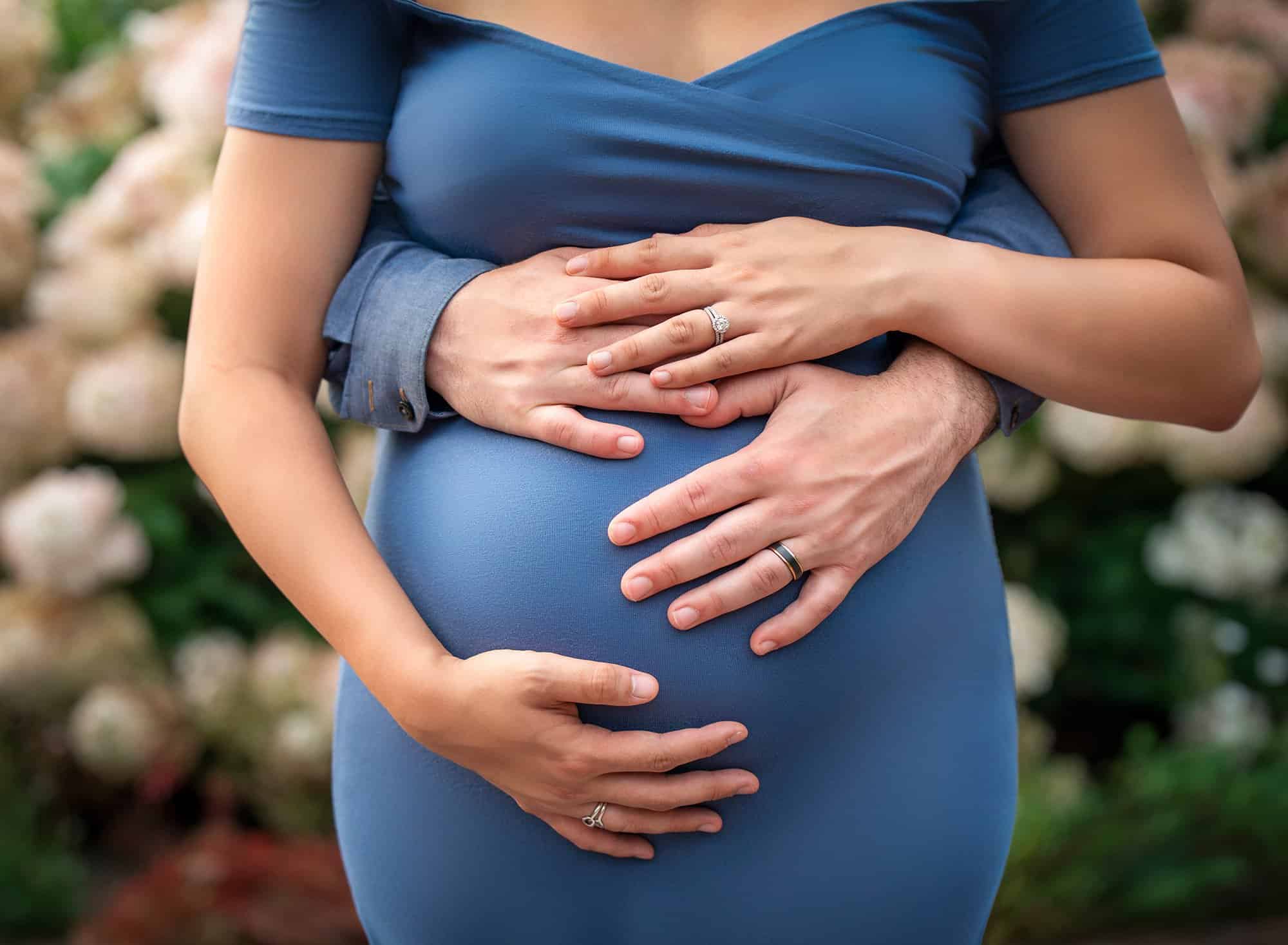 pregnant woman in blue dress shown from the neck down holding her stomach as well as her partners hands