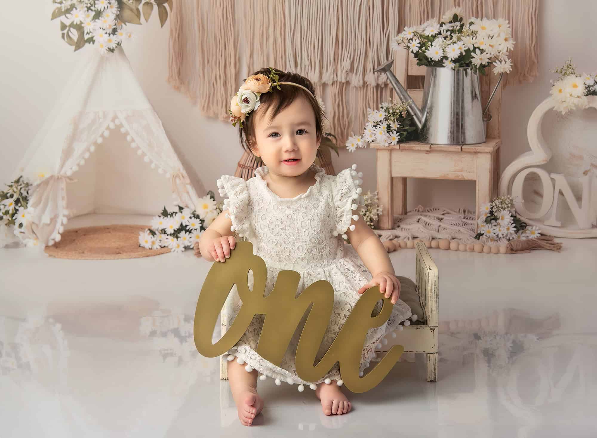 Boho 1 Year Old Photos Sophie sitting on a small, painted bed, holding a gold "ONE" sign