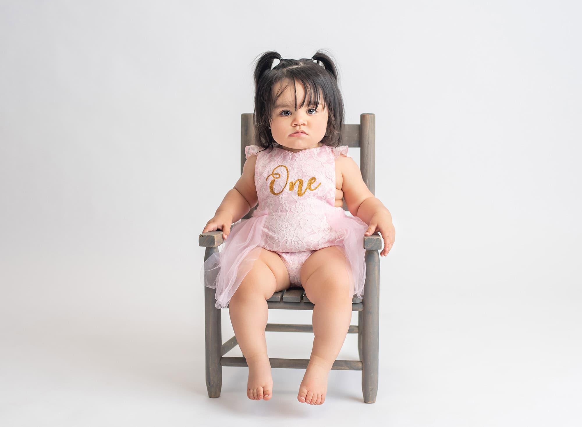 one year old baby girl in pigtails and pink laced one dress sitting on rustic chair with neutral background