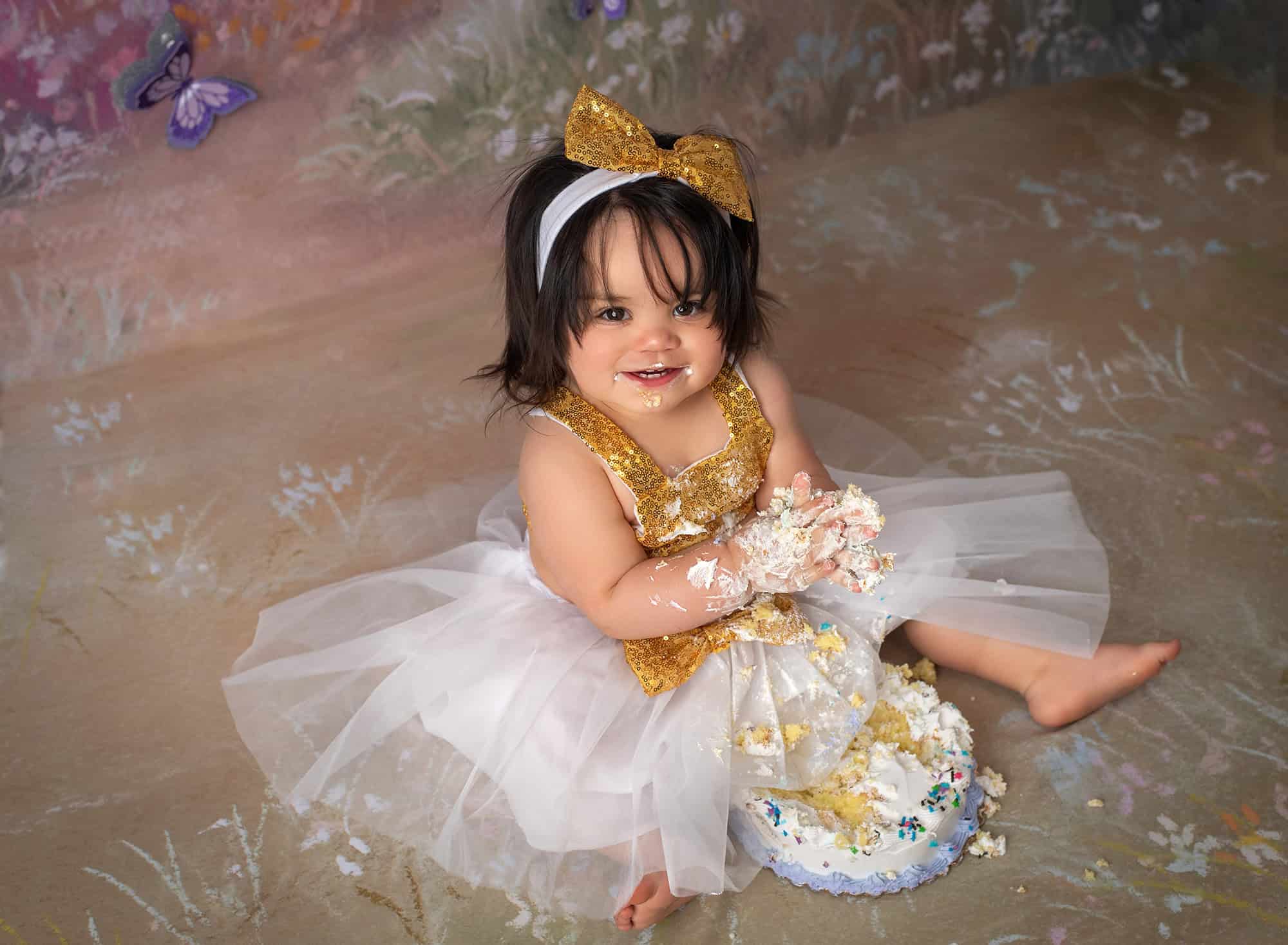 one year old girl dressed in white and gold smashing cake