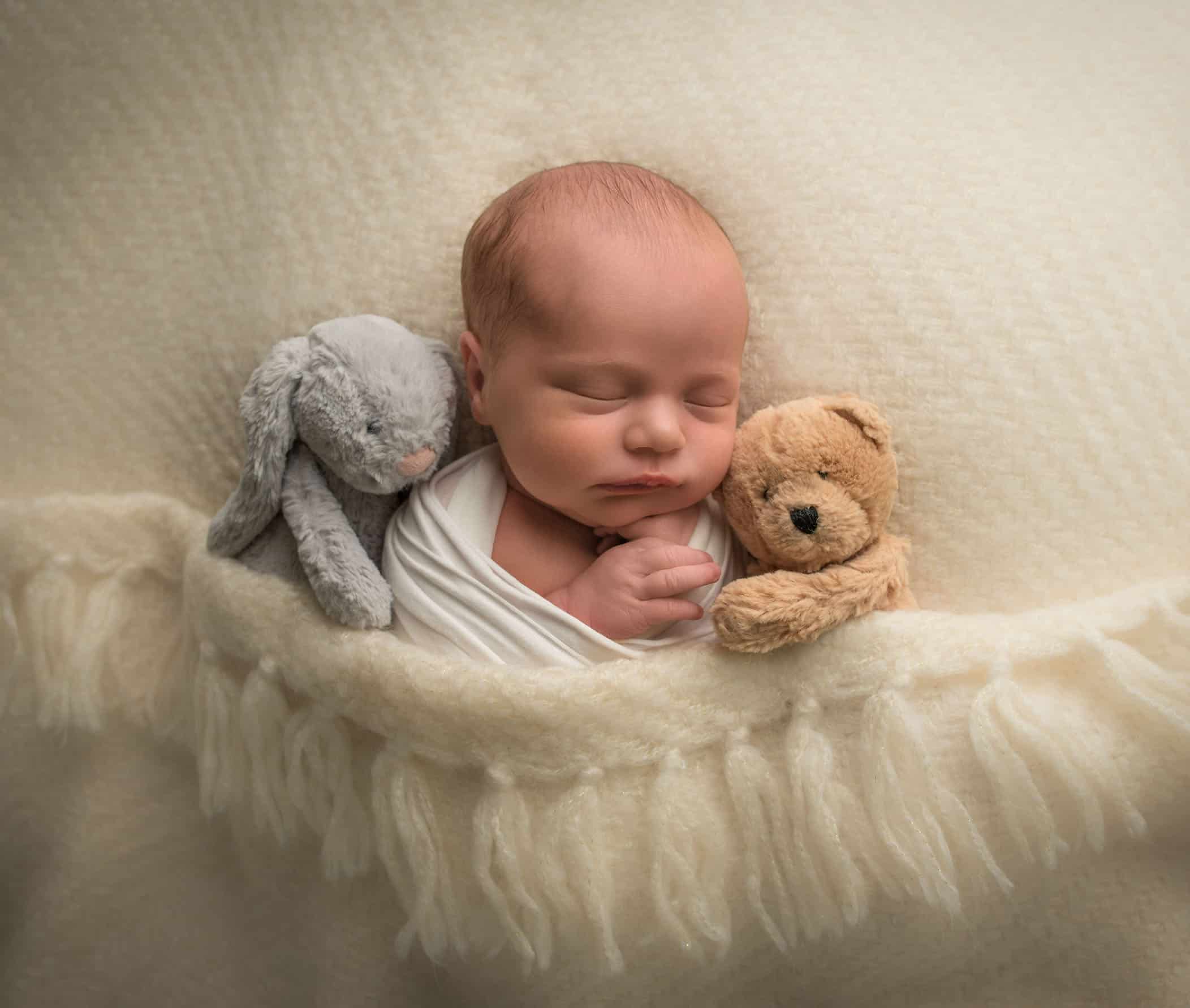 newborn baby girl sleeping on cream blanket with stuffed animals hugging her
