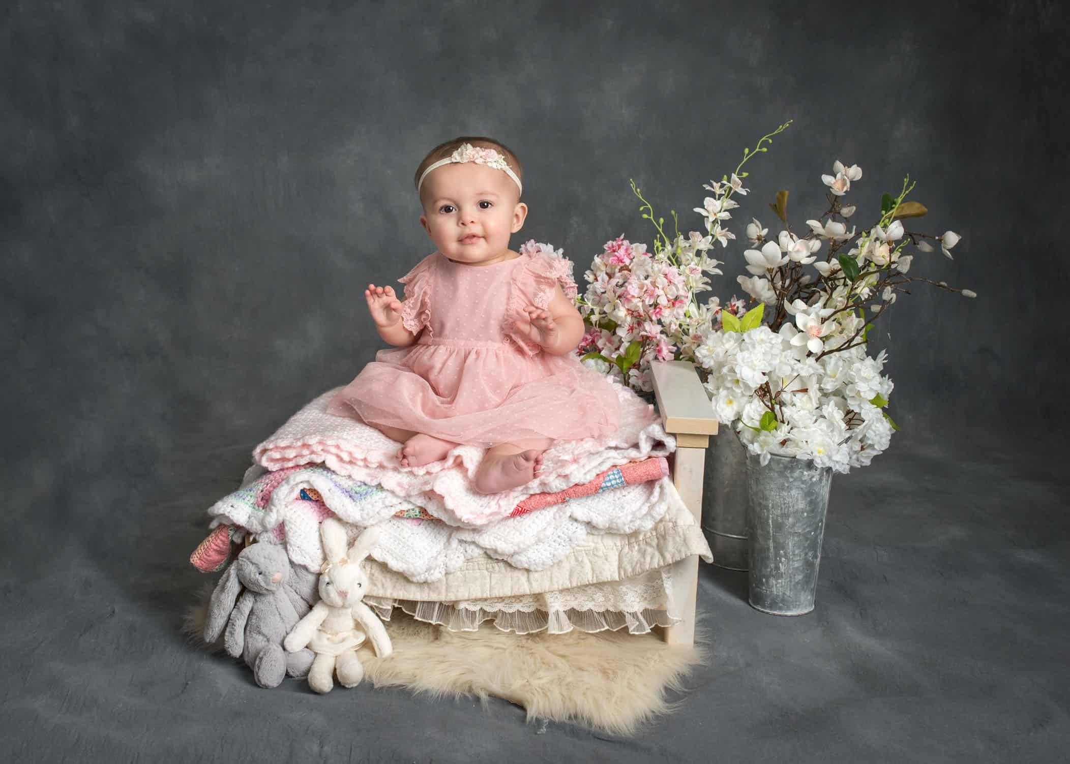6 month old baby girl sitting on doll bed covered in layers of blankets with toy rabbits and flowers