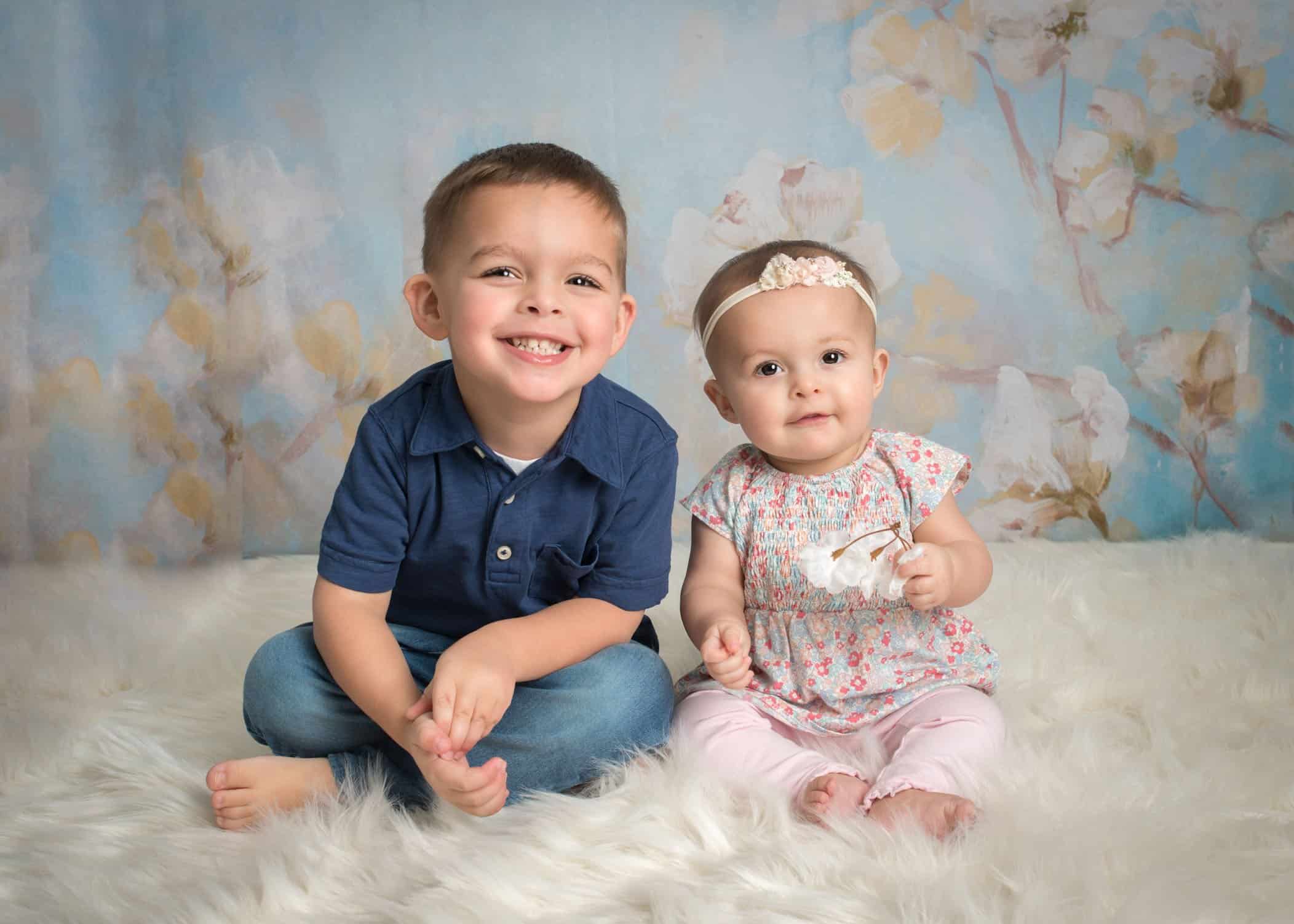 6 month old baby girl sitting with 3 year old brother on white fur smiling
