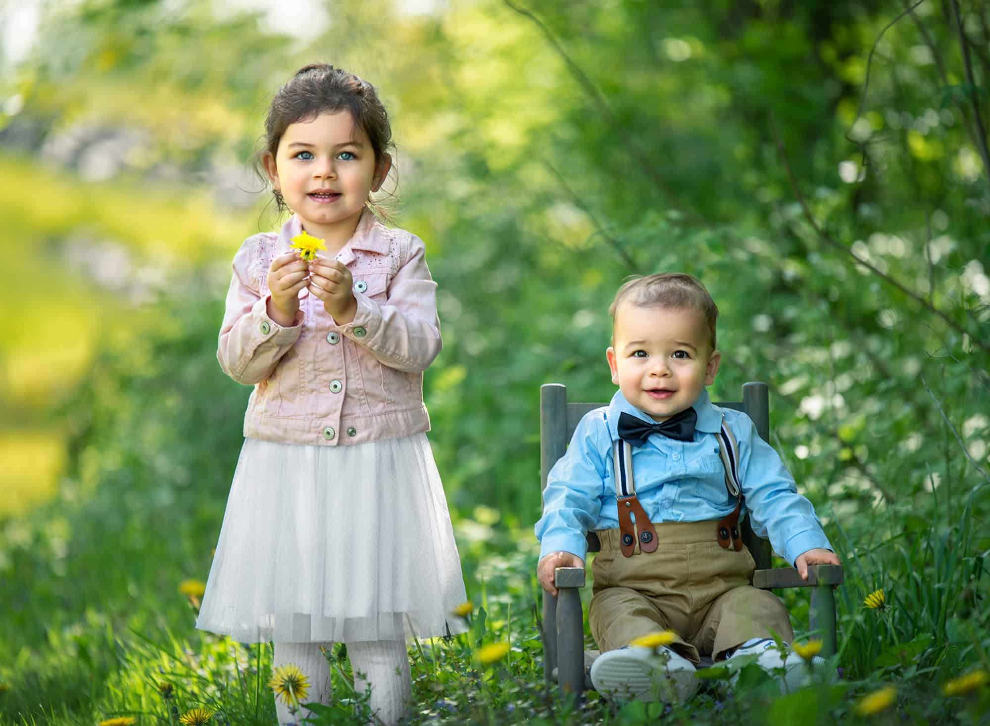 Summer Family Photos young girl holding dandelion standing next to infant brother sitting in a rustic mini chair in a dandelion field