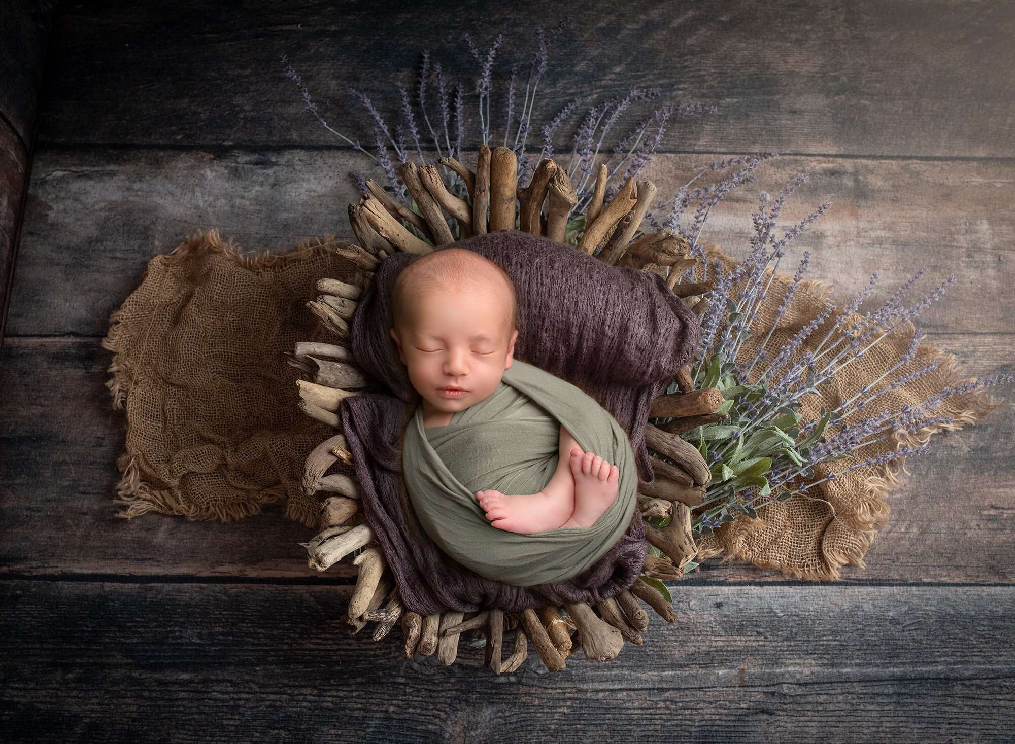 newborn baby boy swaddled in green laying in woodsy basket with lavender sprouting underneath