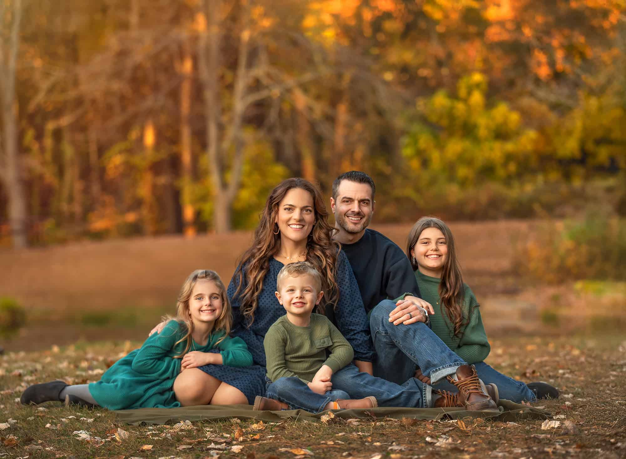 Family of five seated on a blanket in a park with glowing fall foliage.