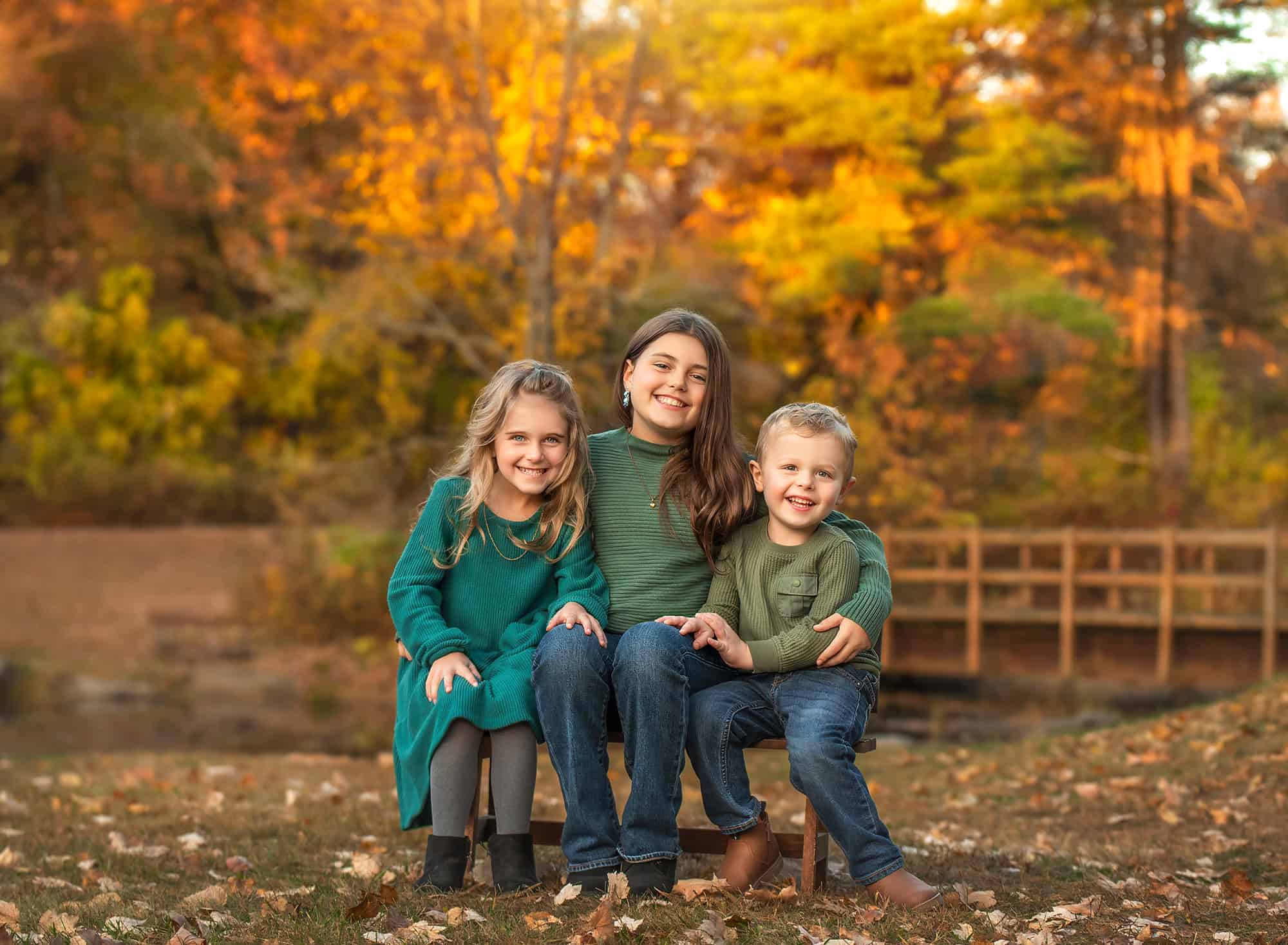 Three siblings seated together on a small bench, vibrant leaves and a footbridge in the background.