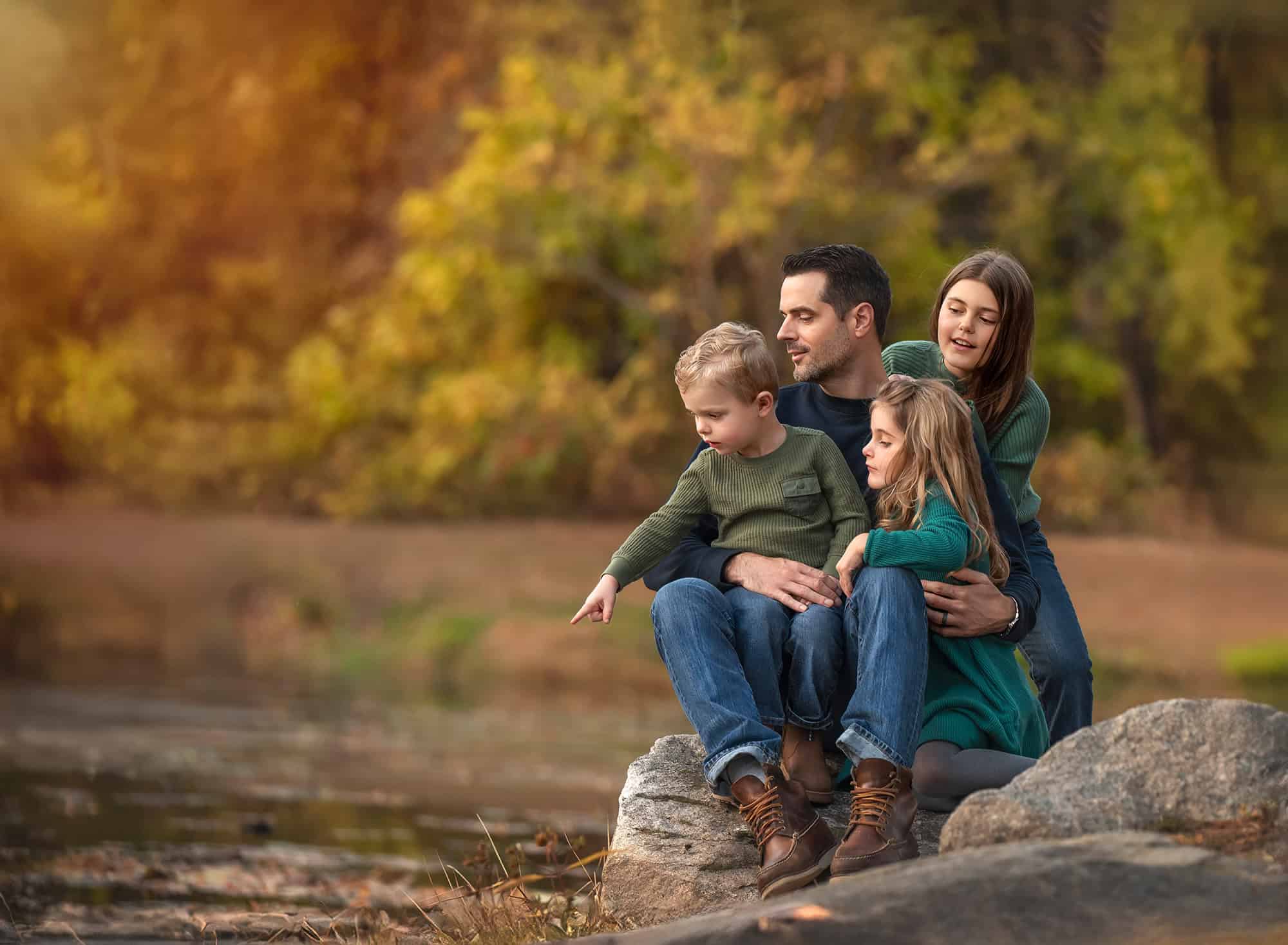Father sitting on rocks by the water with the three children, toddler pointing toward the pond.