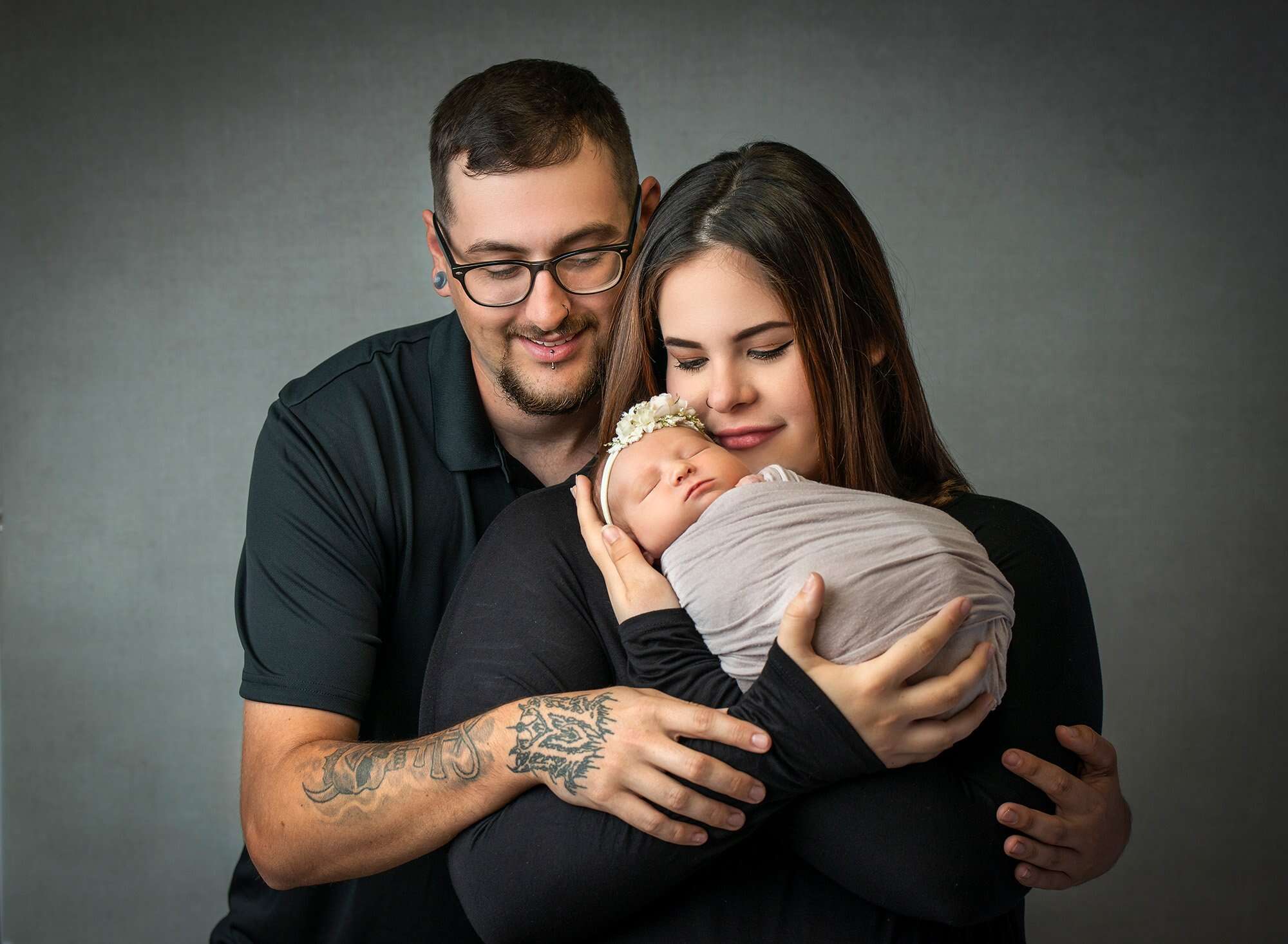 new parents cradling newborn baby girl wearing floral headband swaddled in grey wrap