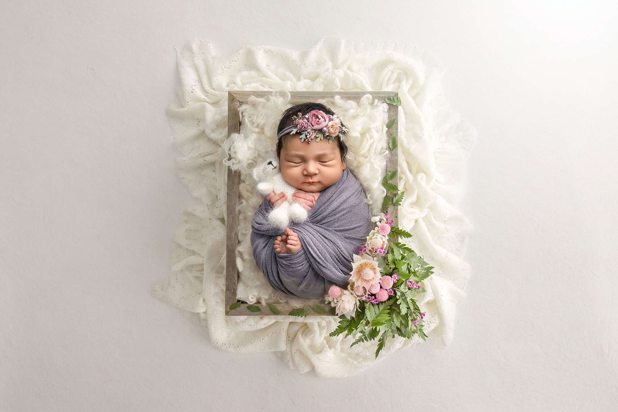 wrapped newborn sleeping in a rectangle crate surrounded by lace and flowers holding a teddy bear