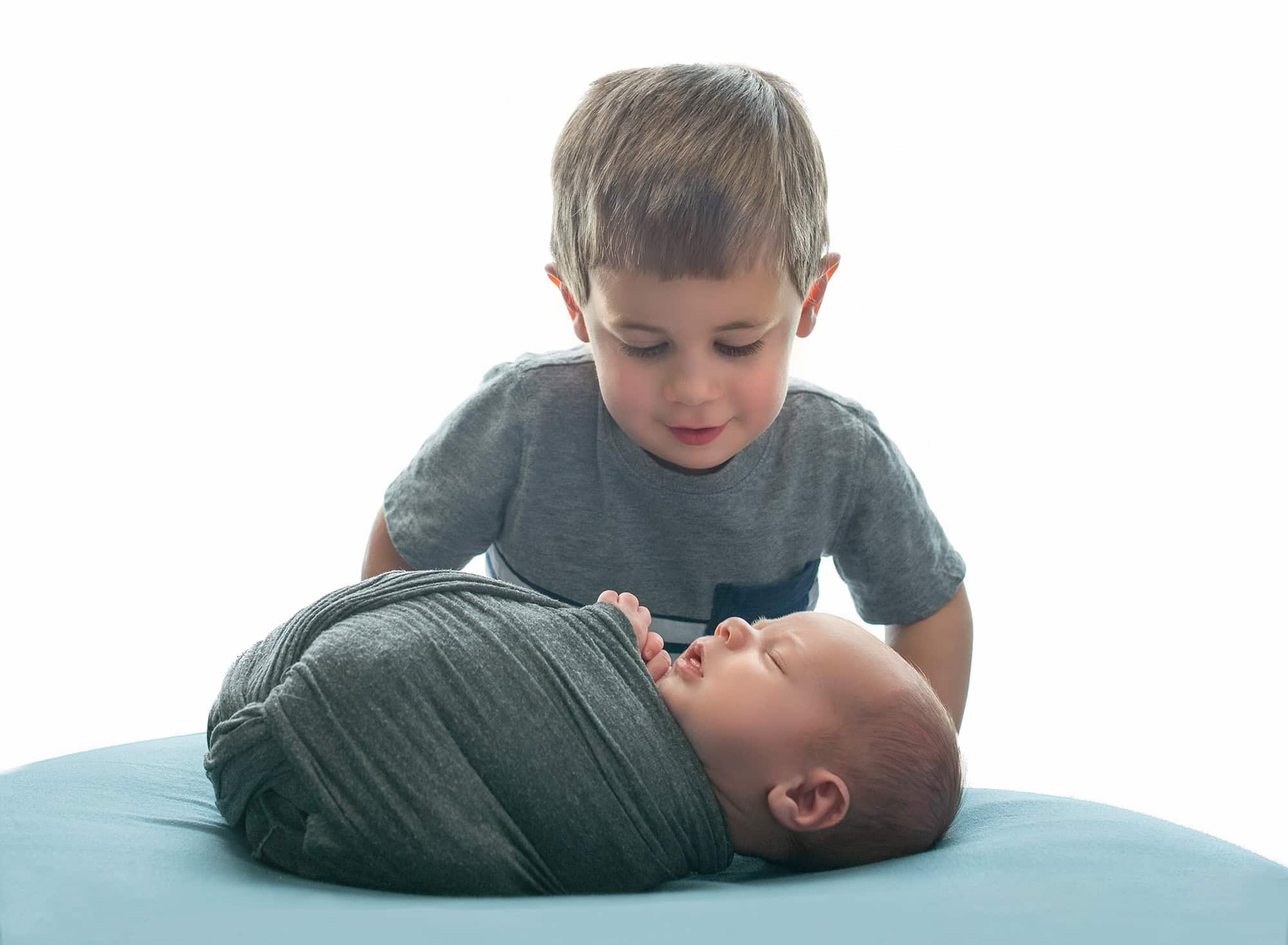 older brother looking down at sleeping newborn baby boy swaddled in gray