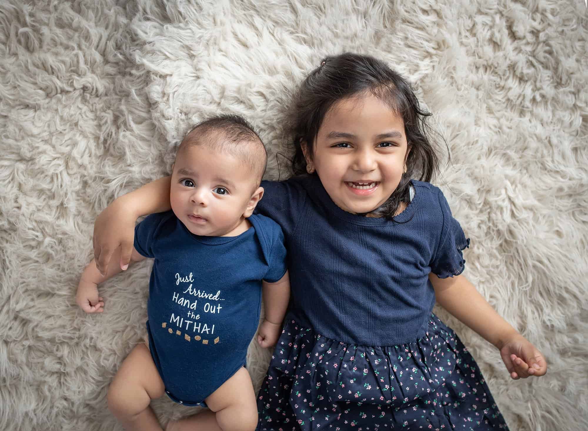 newborn baby boy laying next to older sister both dressed in navy blue on white fuzzy blanket