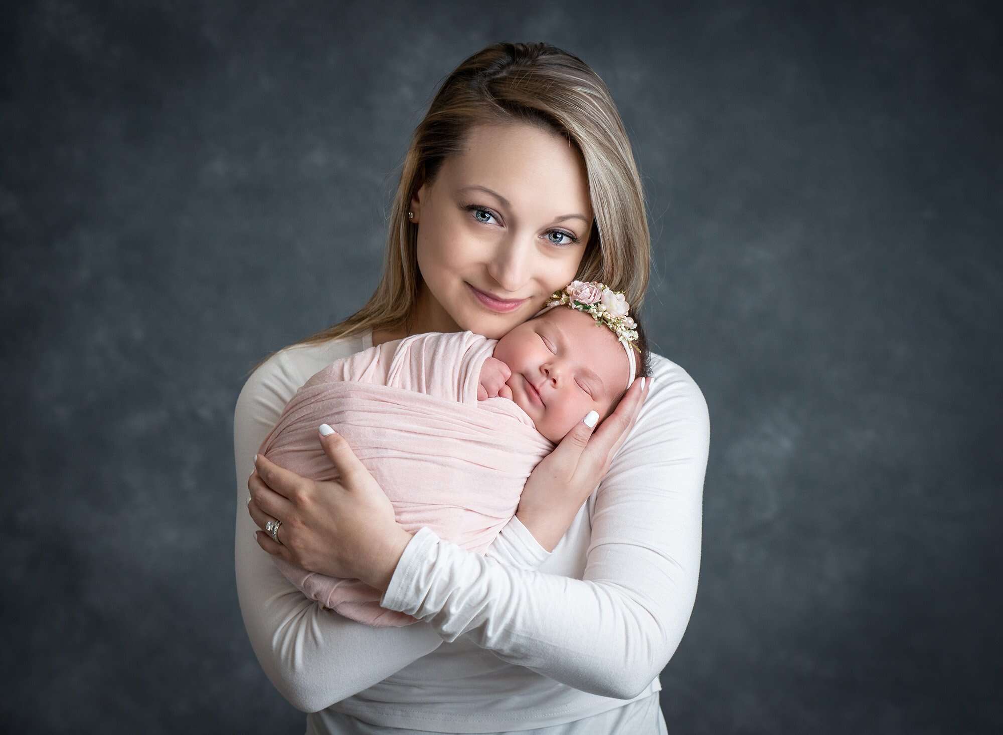 mother cradling newborn baby girl swaddled in soft pink wearing floral headband on dark gray backdrop