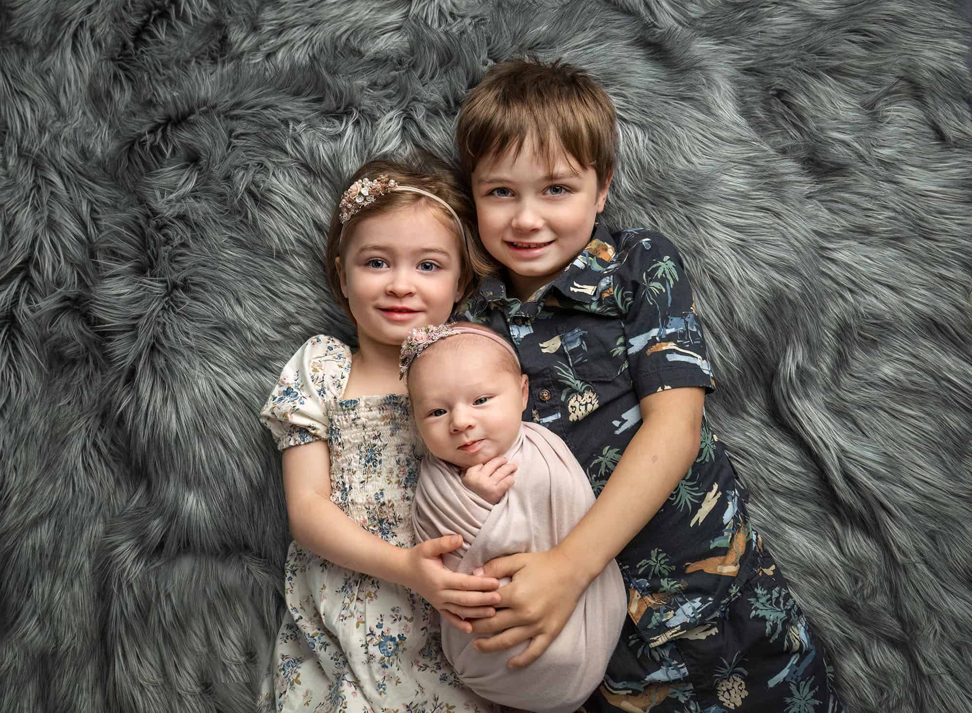 Brother and sister cuddling newborn on gray rug—preparing siblings for newborn photos in studio