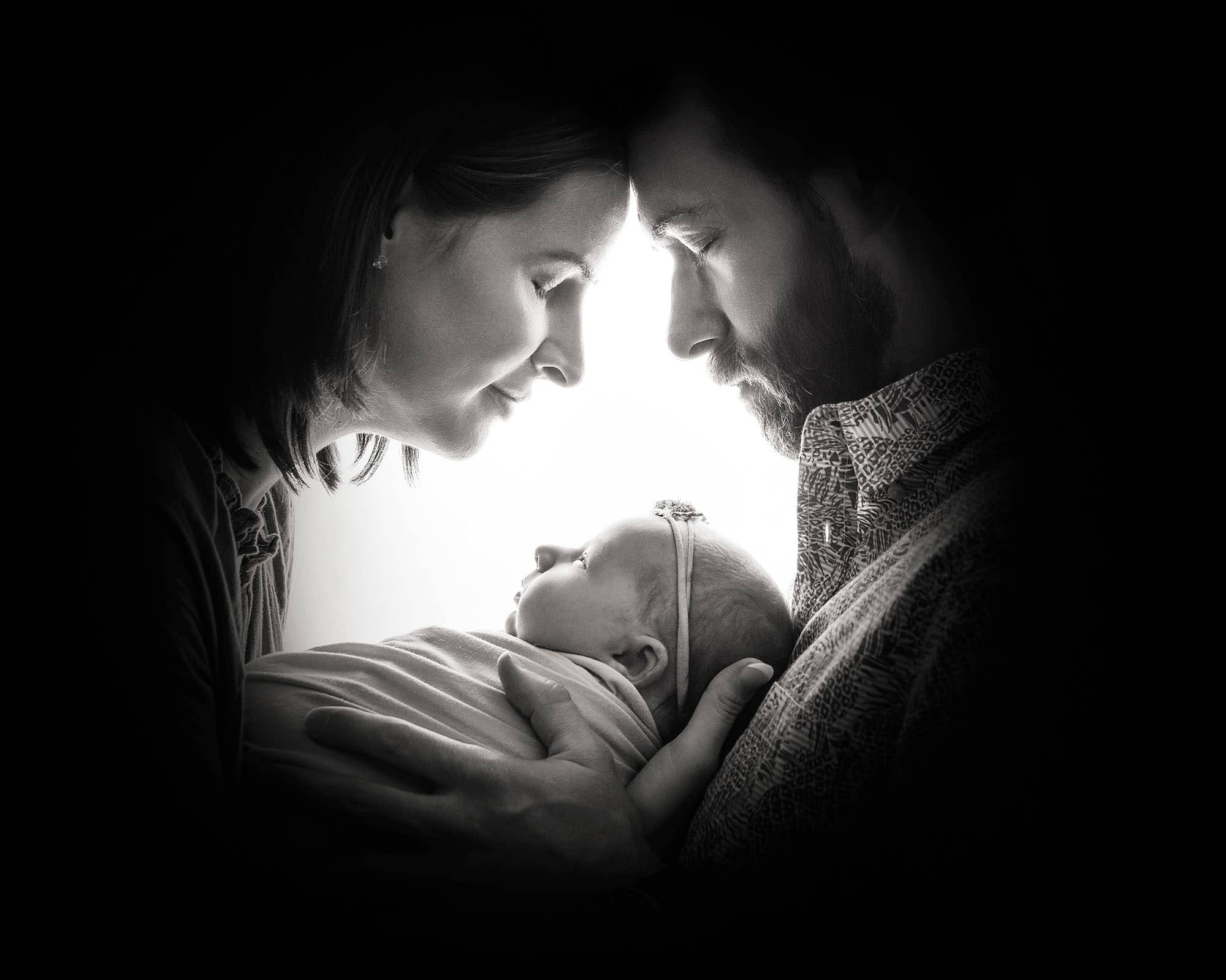 Parents gazing at newborn in backlit portrait—peaceful end to session after preparing siblings