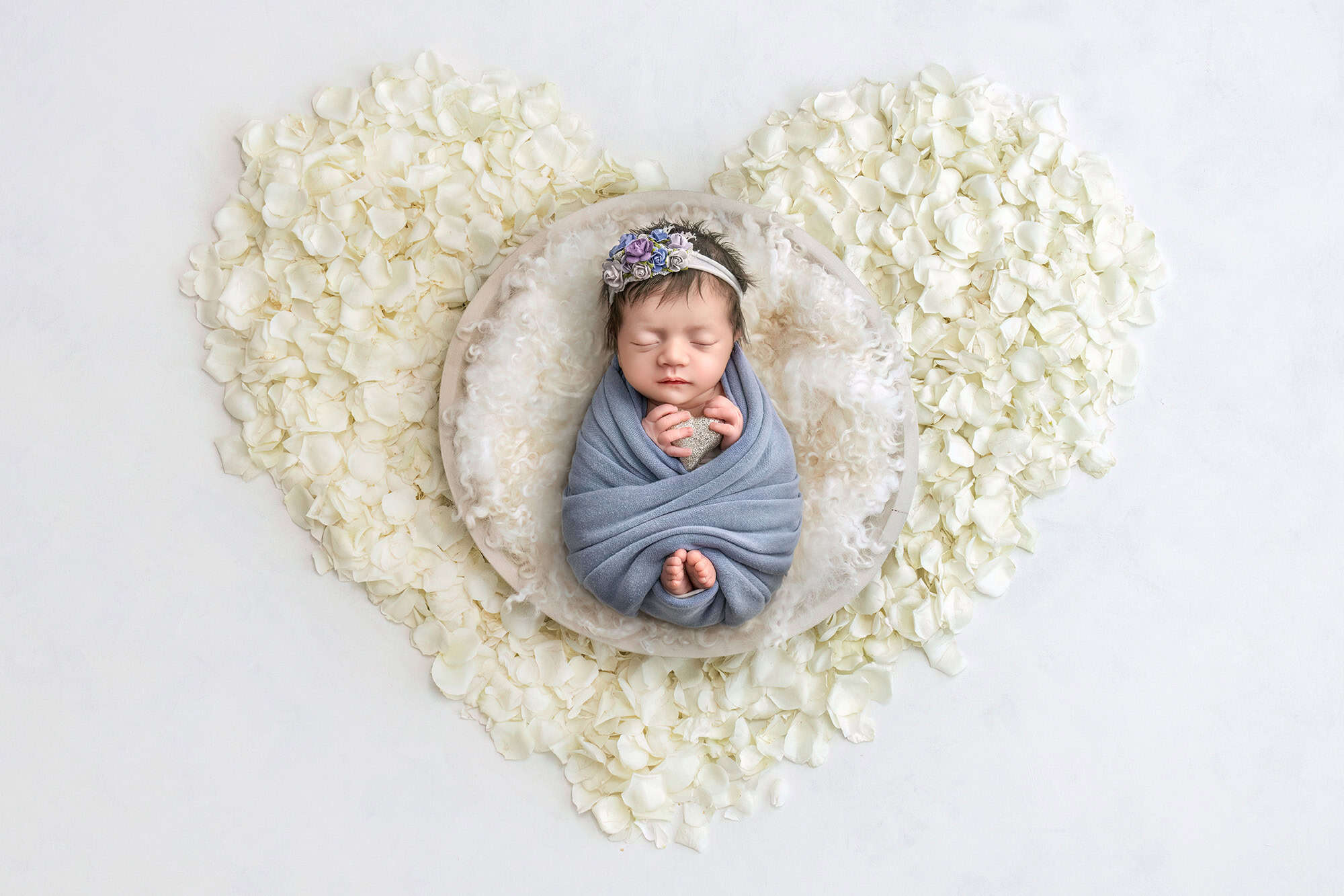 Newborn Lee Anne asleep in a round bowl, wrapped in blue, framed by a heart of white petals.