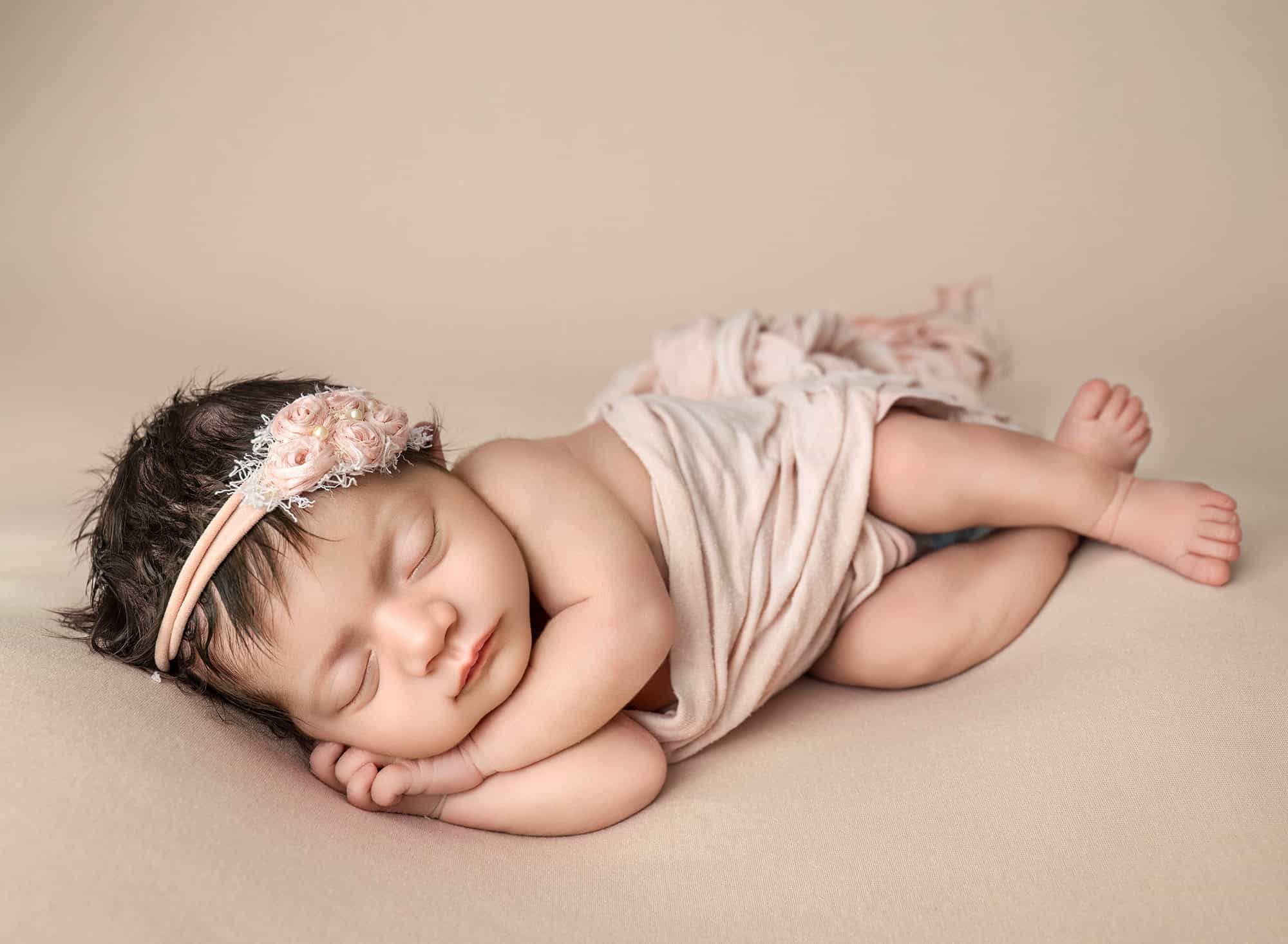 Side-lying newborn pose on beige blanket with soft blush wrap.