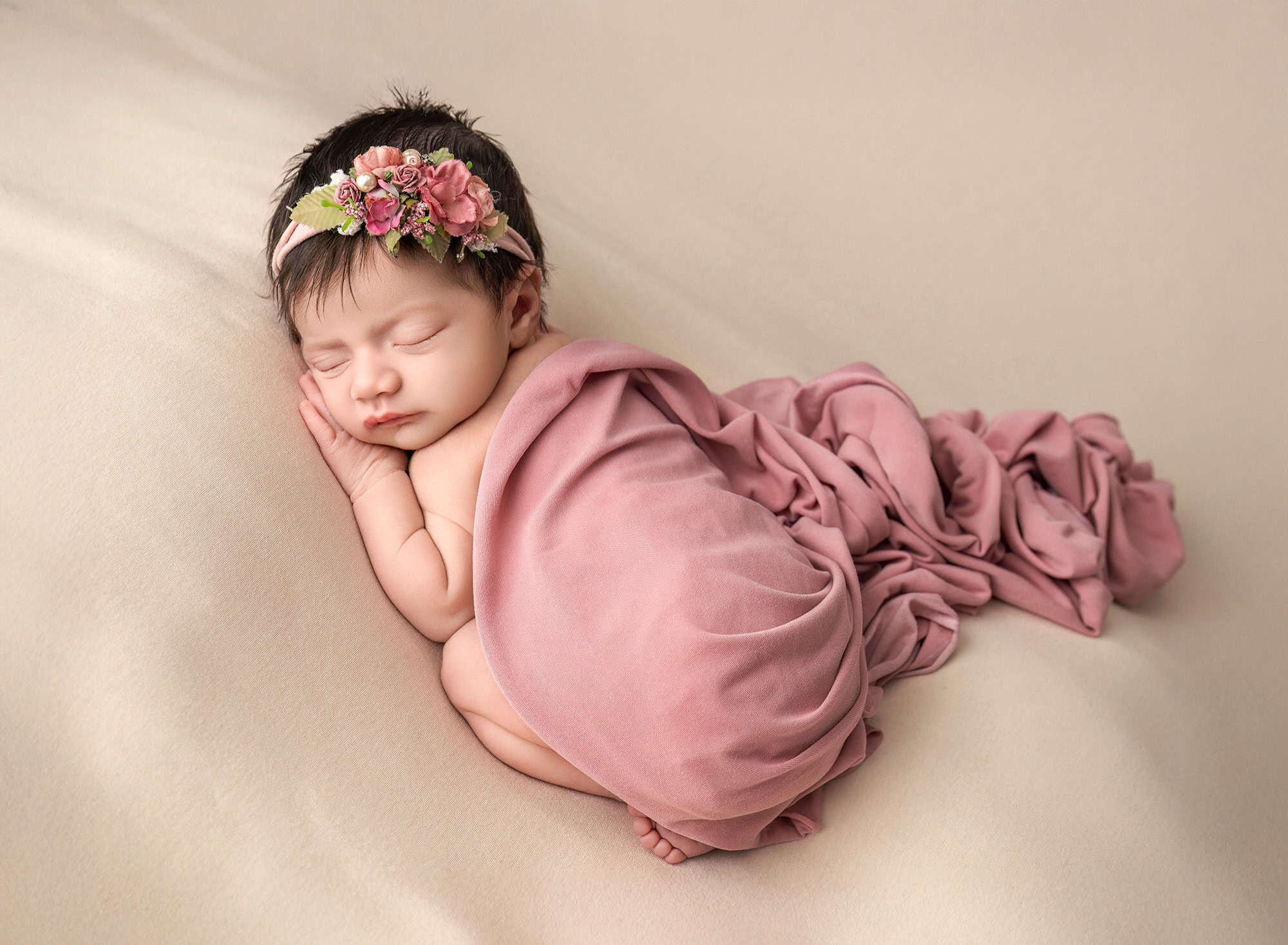tiny newborn baby girl sleeping on a peach fabric with a mauve wrap covering her.