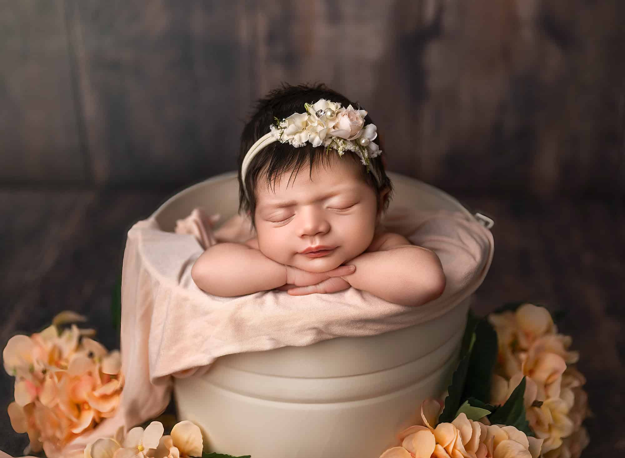 Newborn resting in a cream bucket with peach hydrangeas at the base.