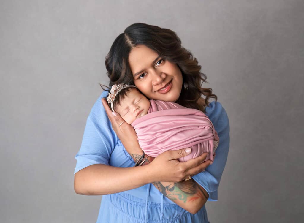 Mother in blue dress holding sleeping newborn wrapped in pink.