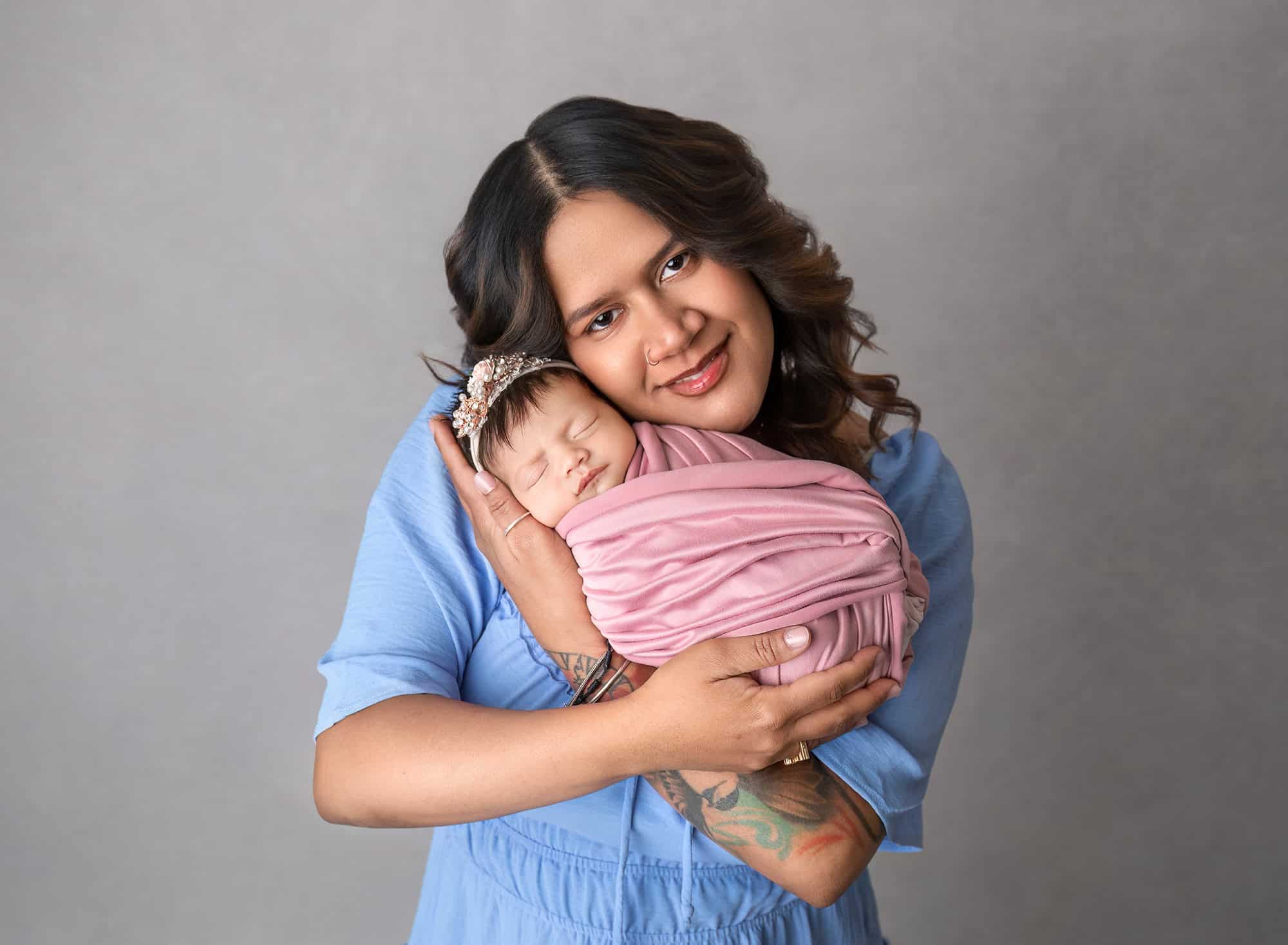 Mother in blue dress holding sleeping newborn wrapped in pink.