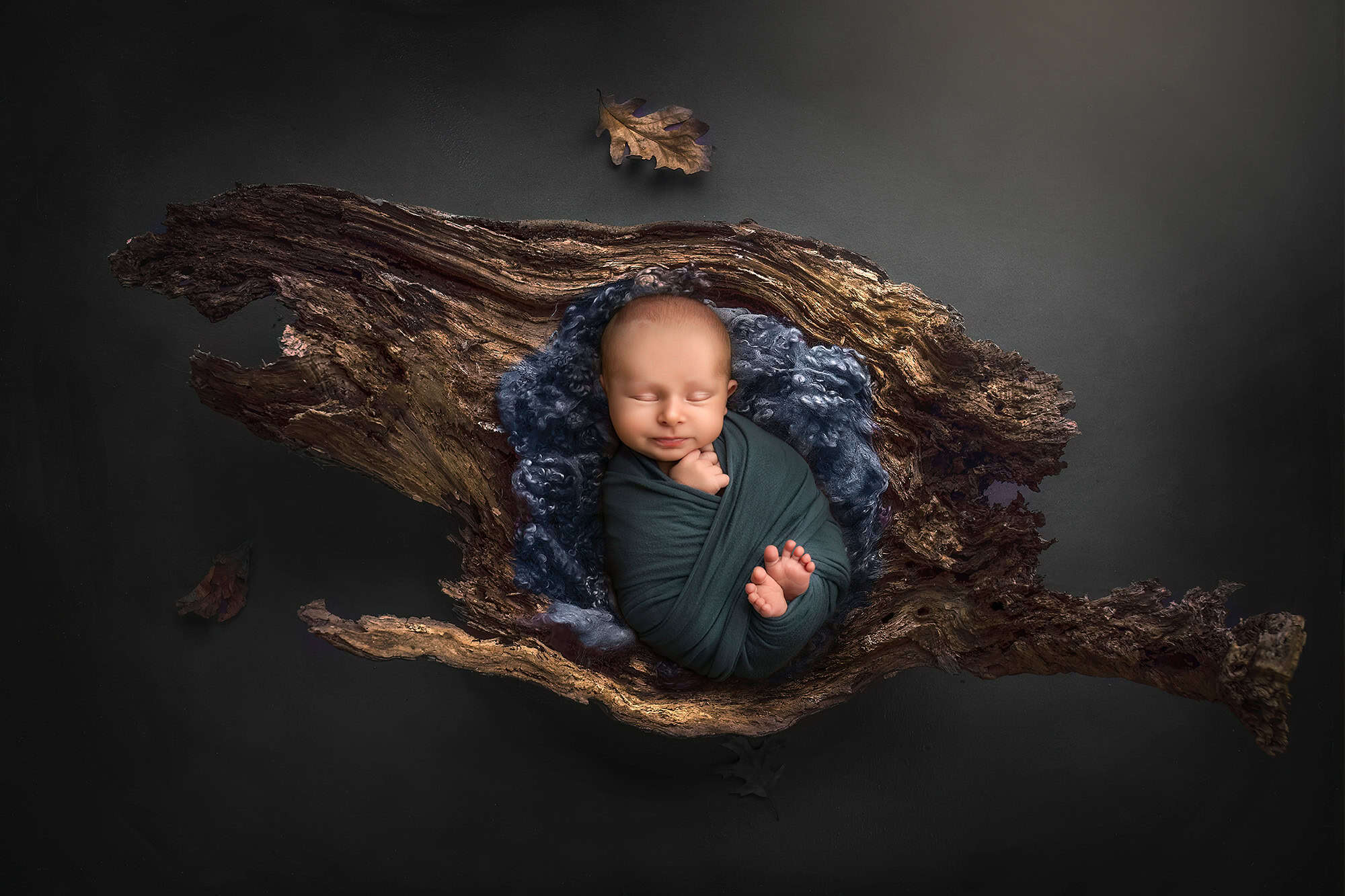 Sleeping newborn Levi in deep green wrap, resting in a natural wood prop with curly wool.