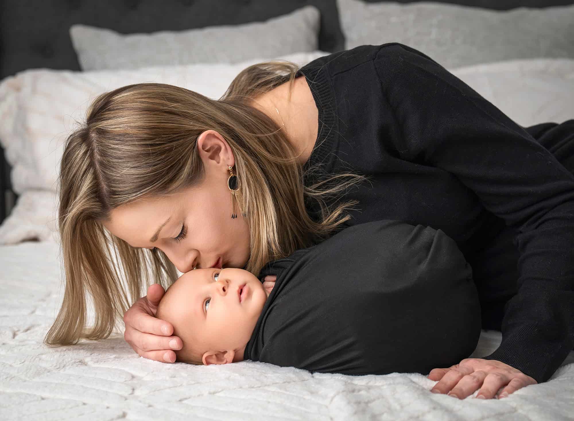 Mother kissing awake newborn Levi on a bed, soft natural light.