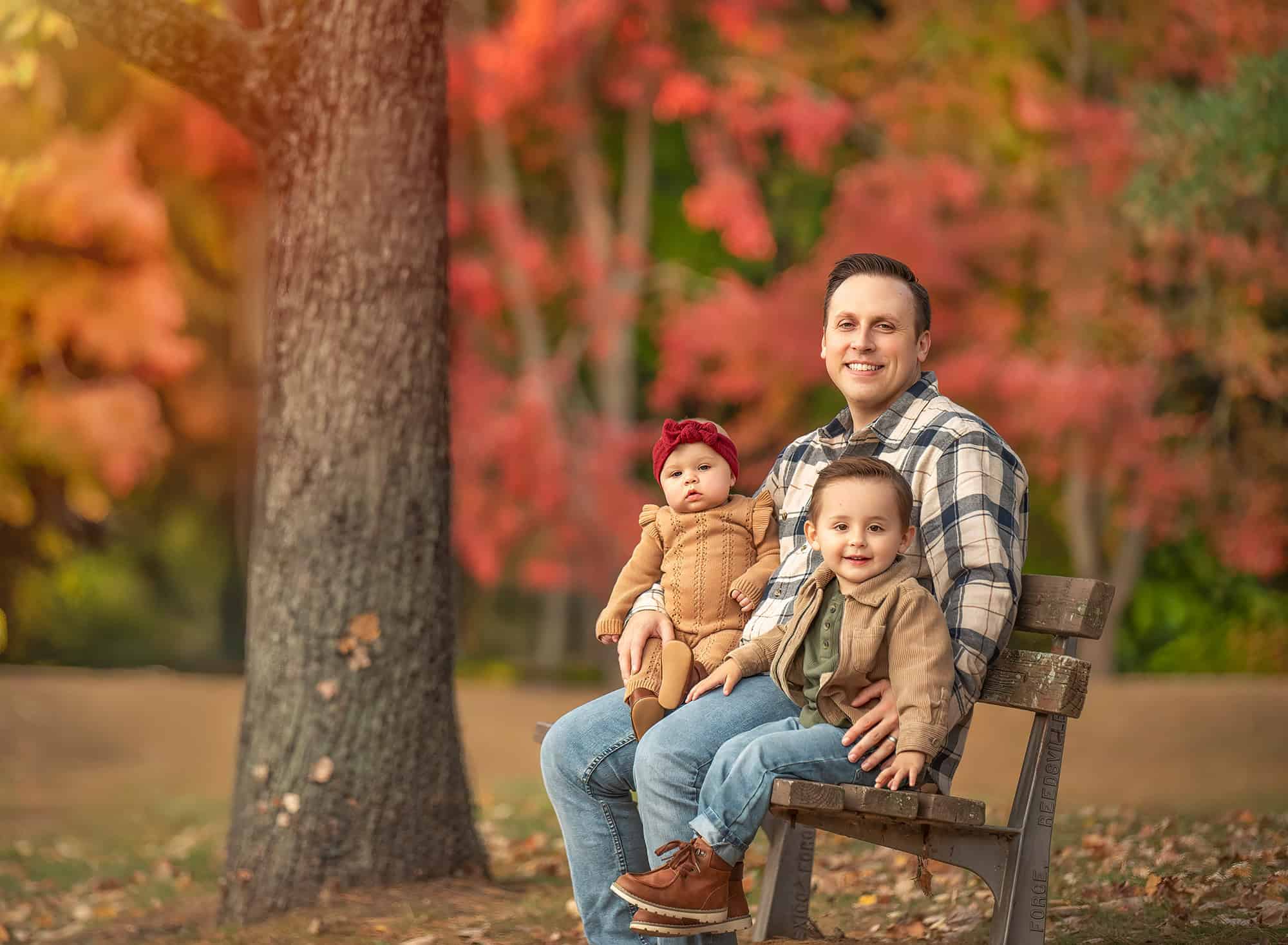 Dad on a park bench with baby Kennedy and toddler Lincoln in fall light.