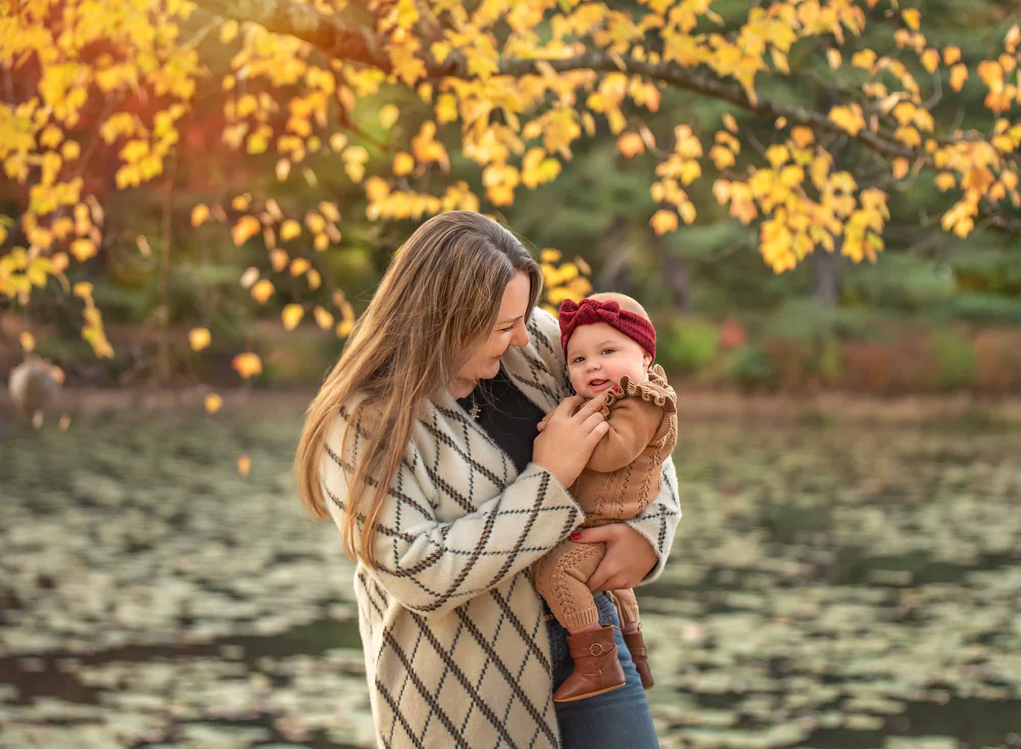 Mom holding smiling six-month-old by a lake with golden leaves.