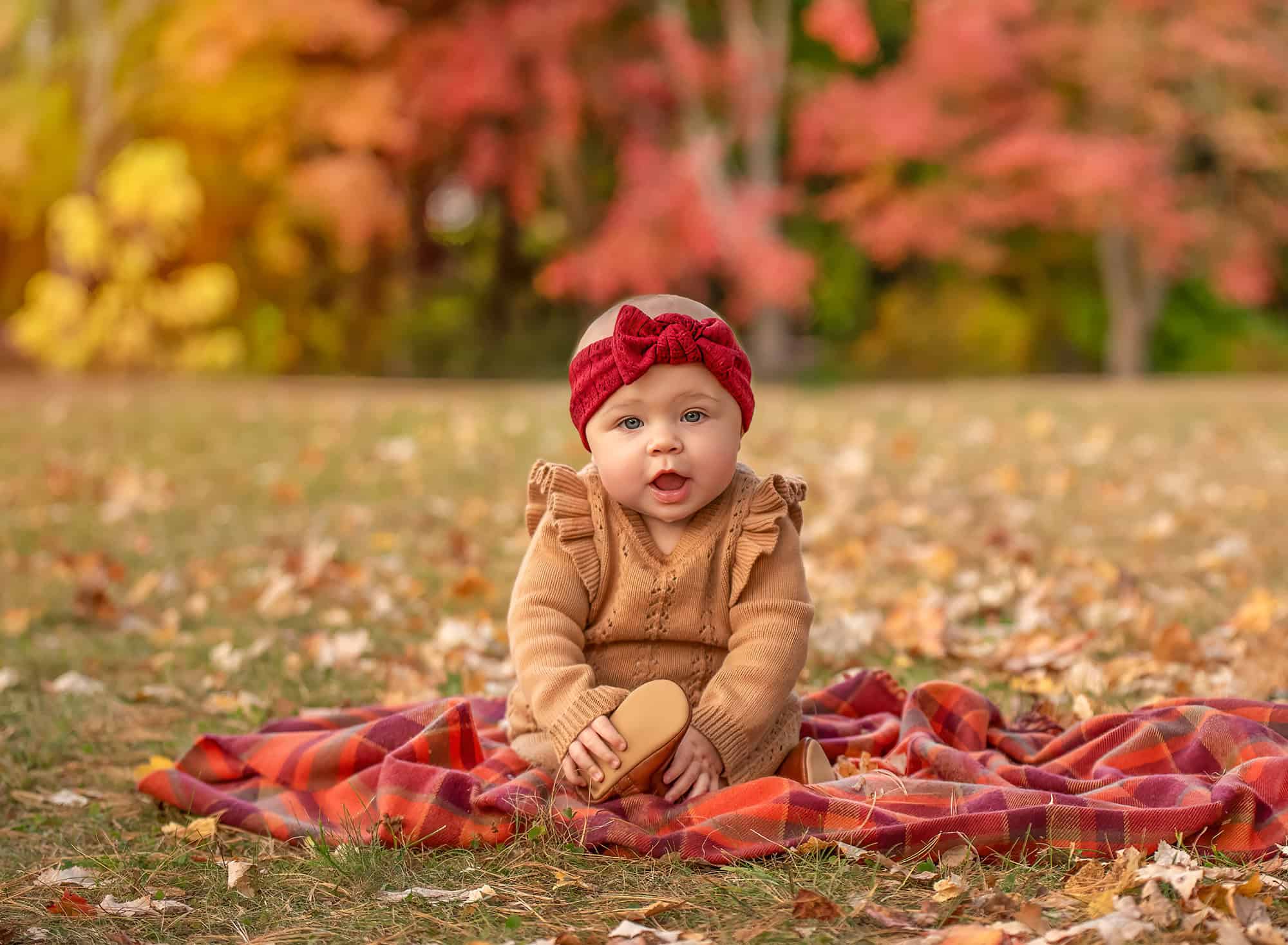 Kennedy sitting on a plaid blanket, smiling with red and gold trees behind.