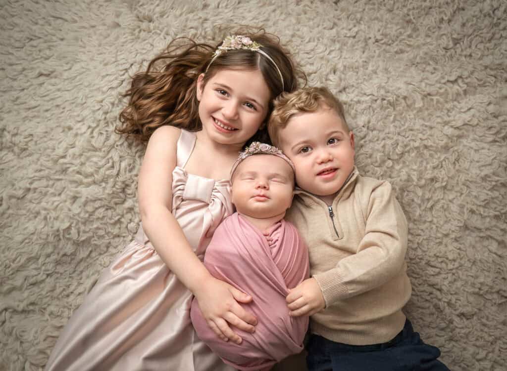 Siblings cuddling newborn on cream flokati — Charlotte and Mason hold sleeping baby Mia in soft neutral tones.