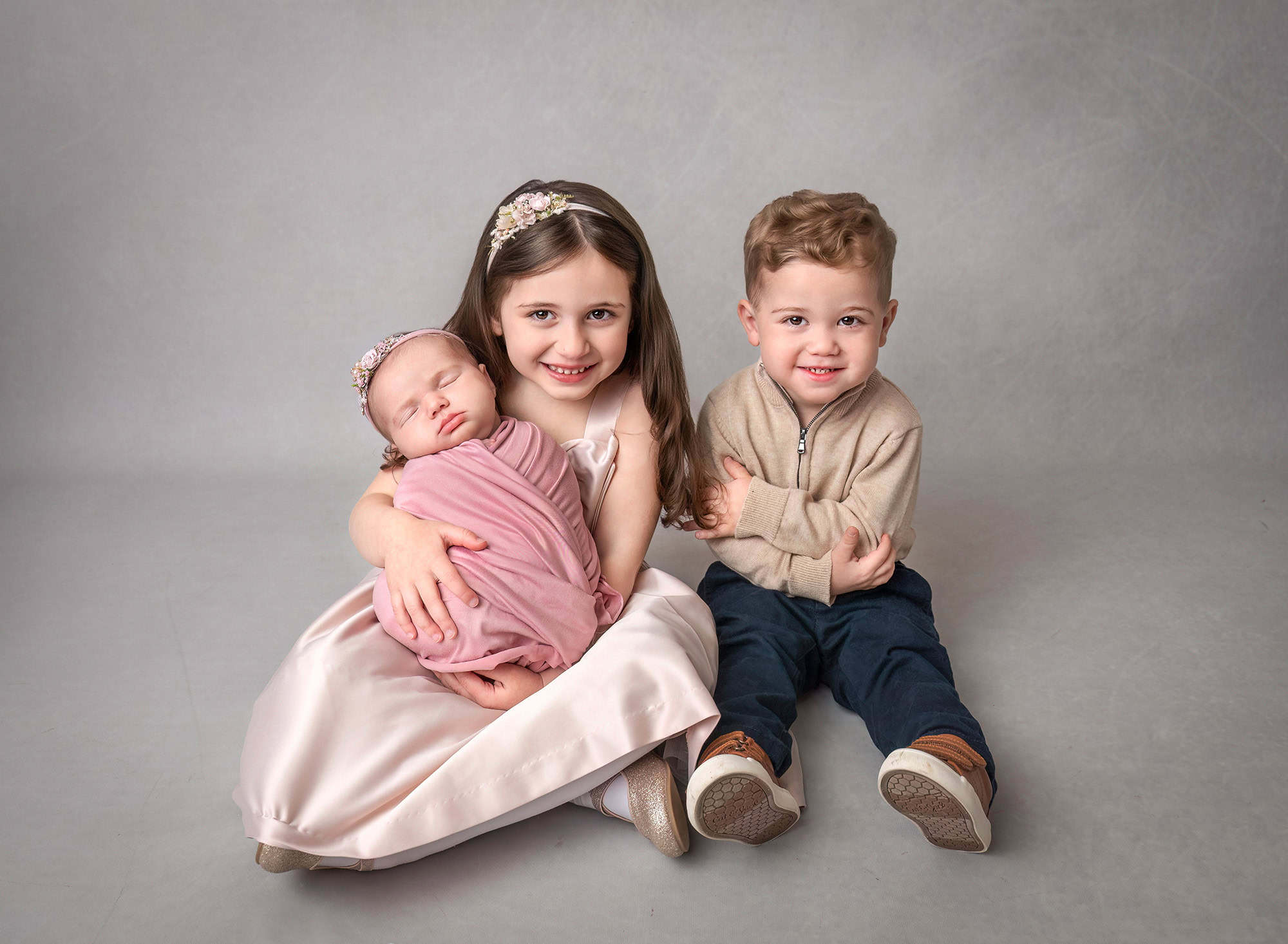 Studio portrait of three children — Big sister hugging newborn while brother smiles beside them.