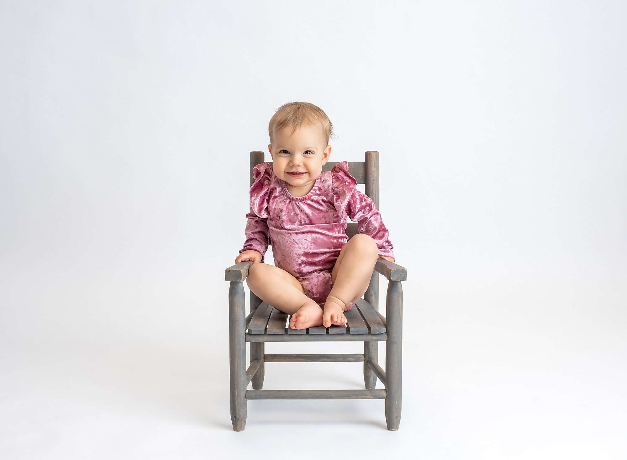 blonde one year old girl wearing pink velvet body suit sitting on rustic chair with white backdrop