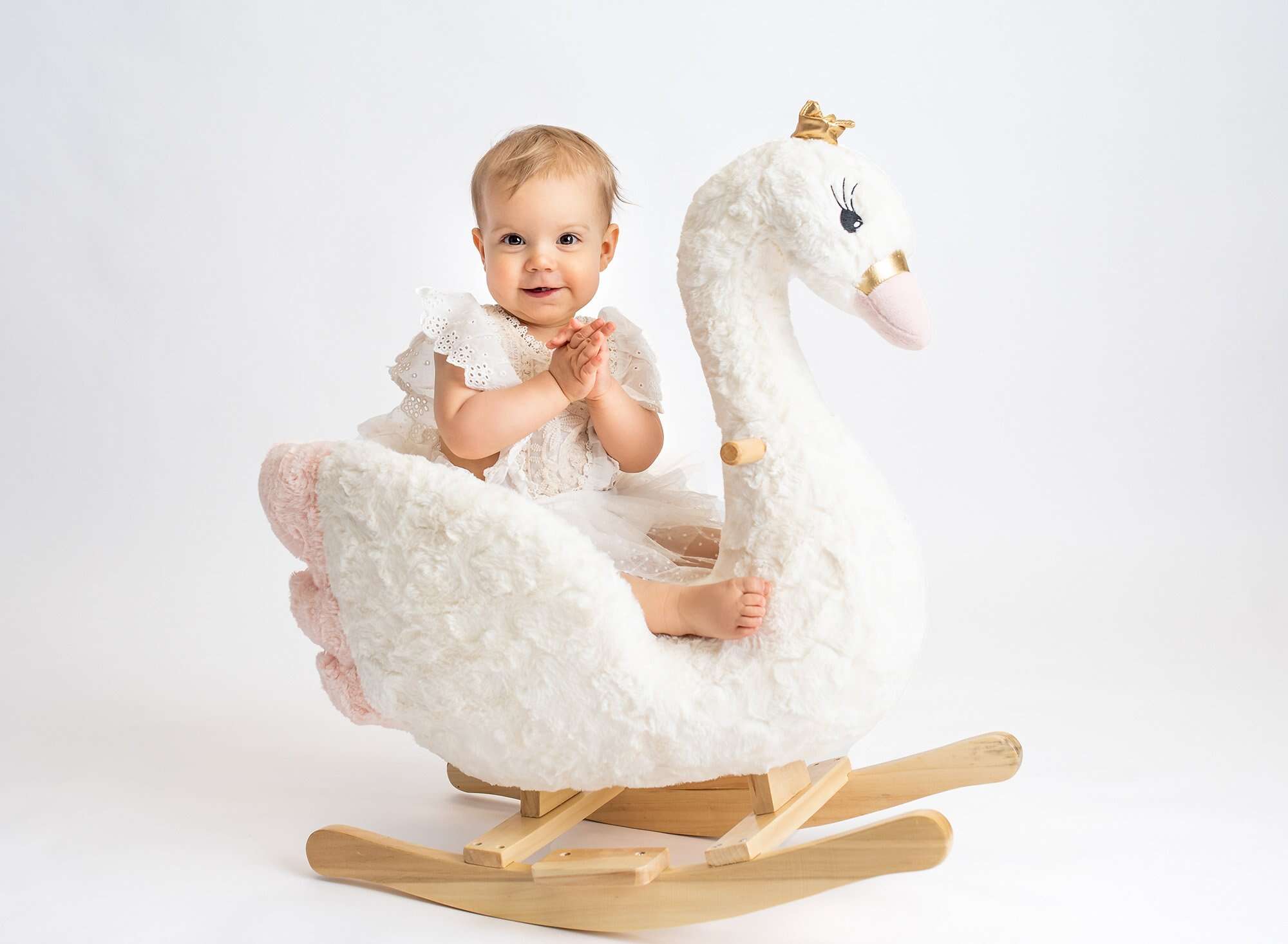 1 year baby photoshoot one year old girl wearing white lace dress sitting on swan rocker with white backdrop