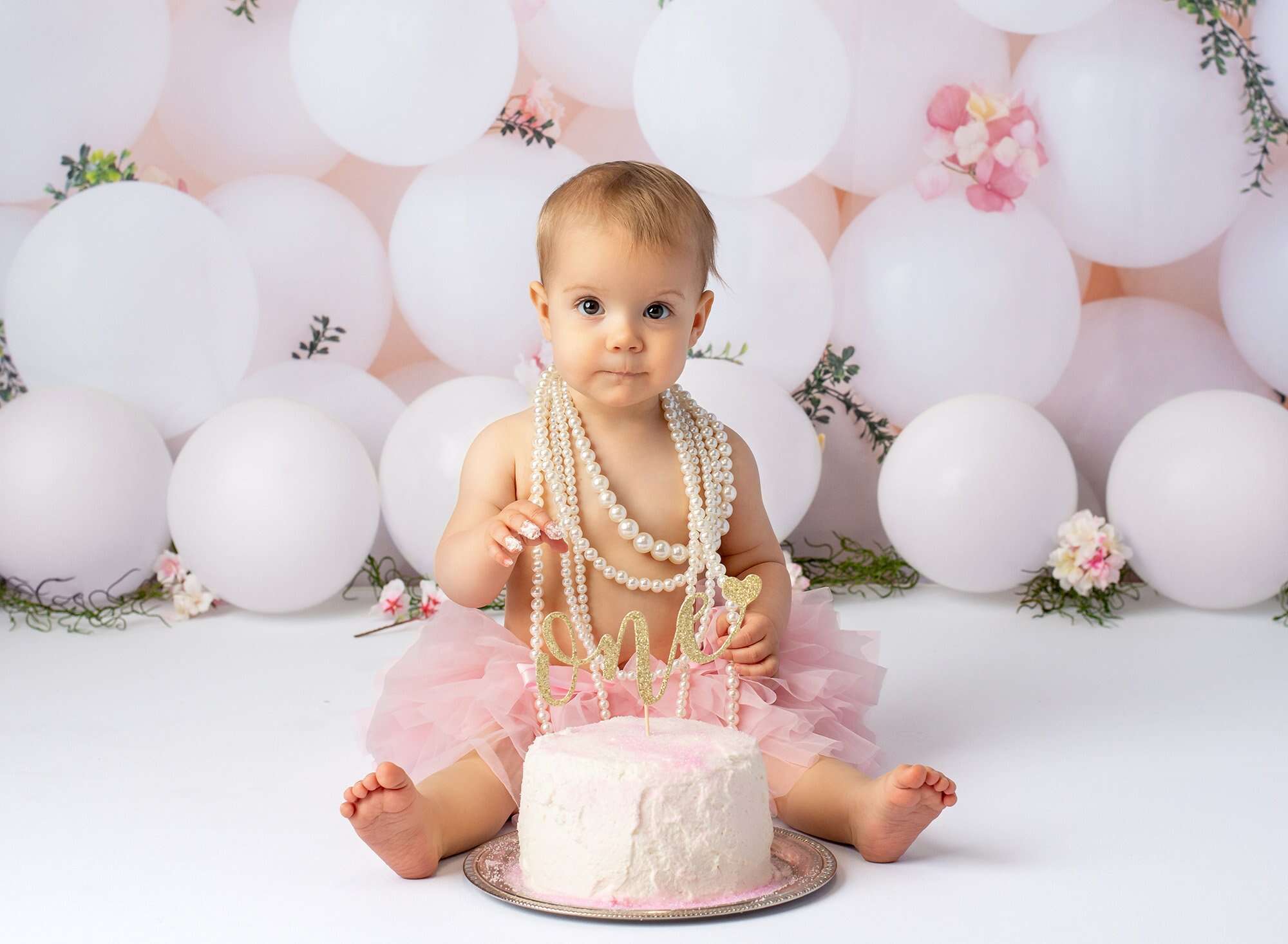 baby girl wearing layered pearl necklace and pink tutu sitting in front of cake with gold glitter ONE sign with white balloons in background and frosting on fingers